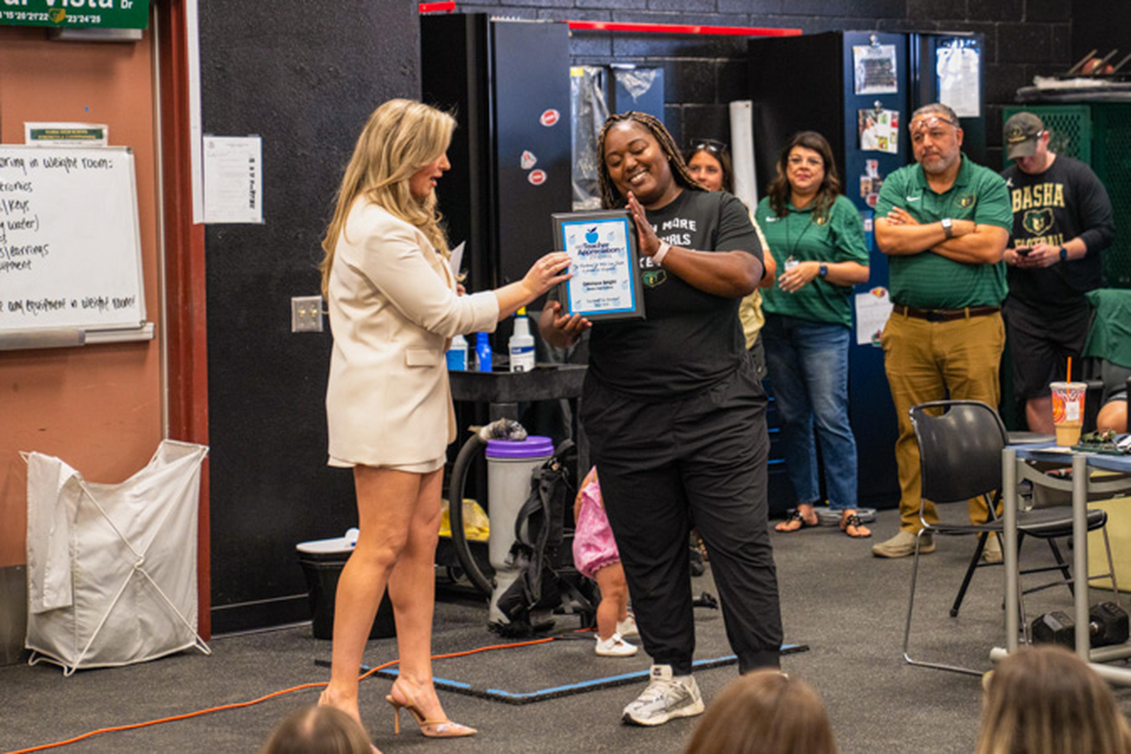 Jen presents an award to Coach Bright, her blazer contrasting her black t‑shirt as students and teachers gather around them in a lively classroom setting.