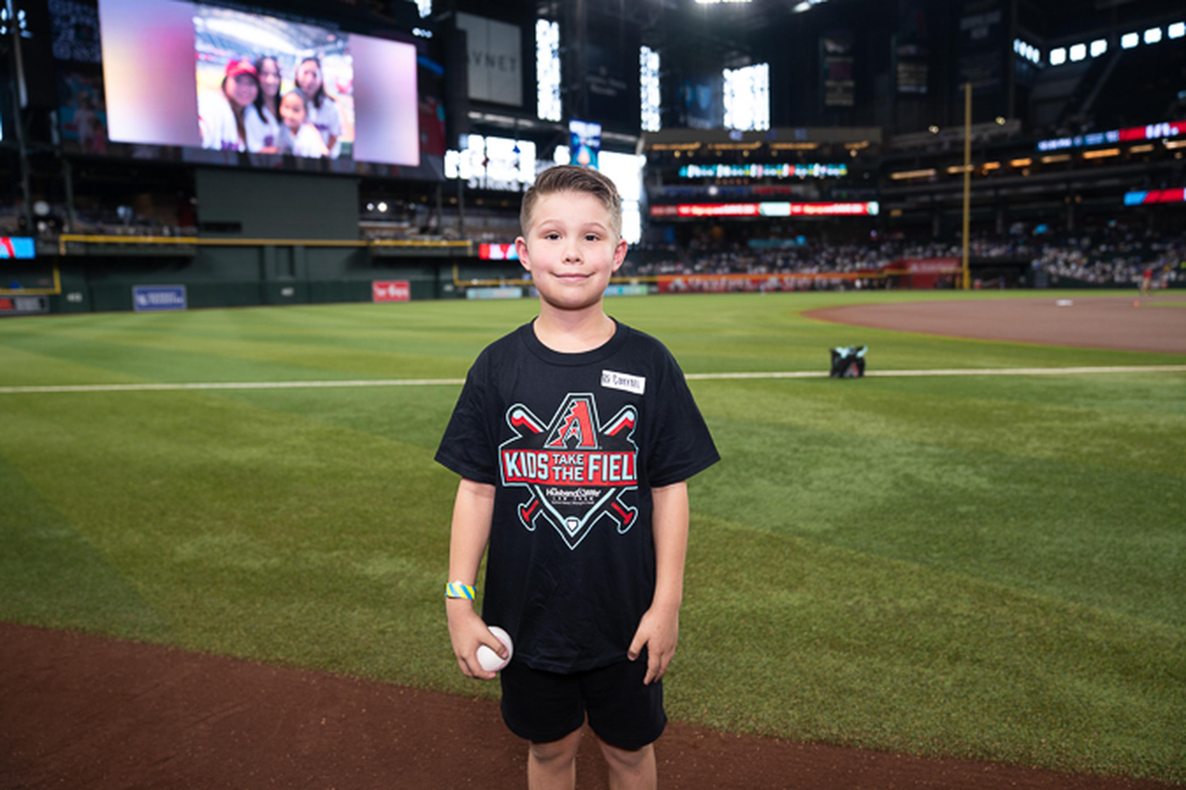 A young Kids Take the Field participant proudly holds a baseball on the field, enjoying an unforgettable experience at the ballpark with the stadium crowd cheering in the background.