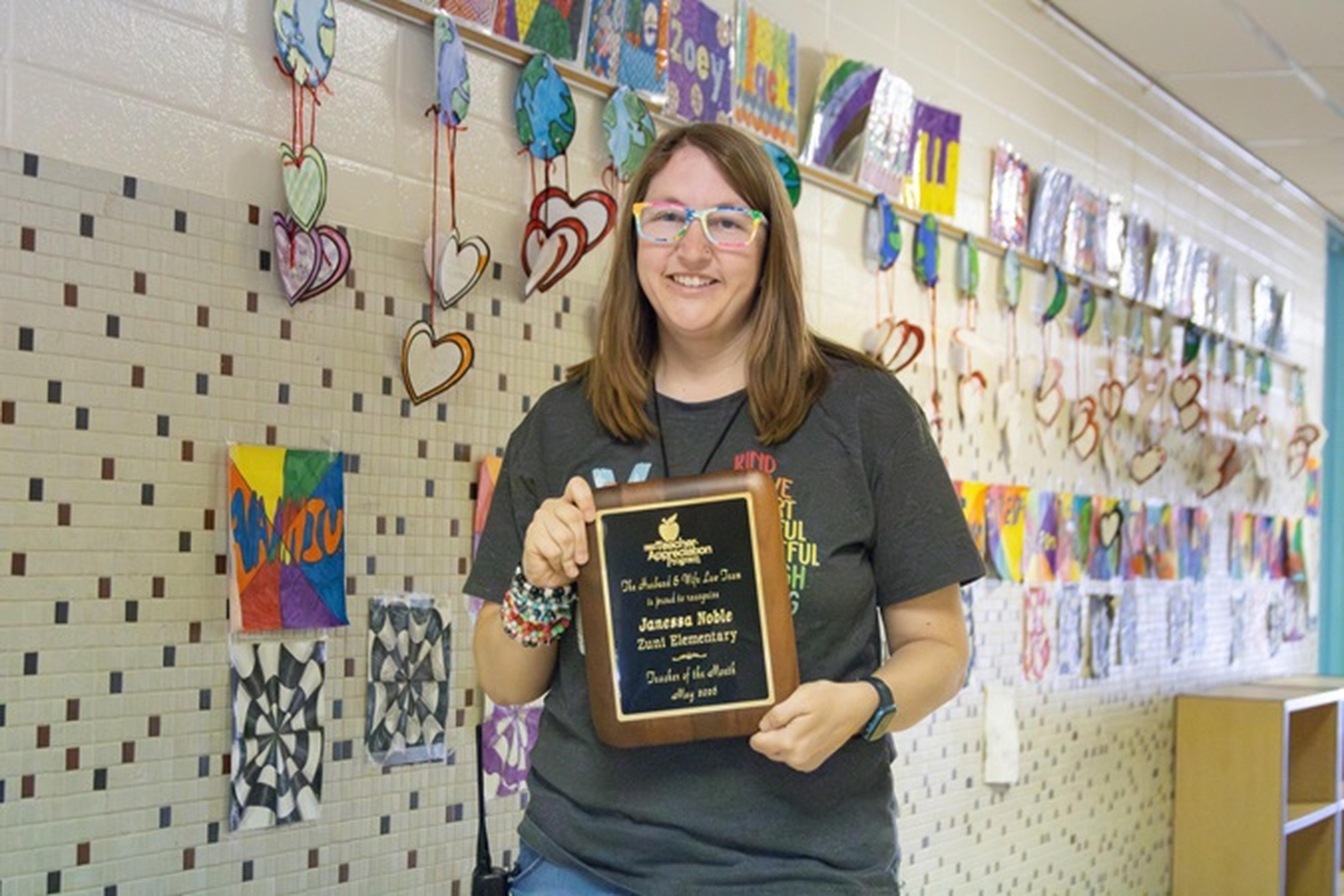 Ms. Noble smiles in a hallway, holding a plaque as artwork lines the walls.