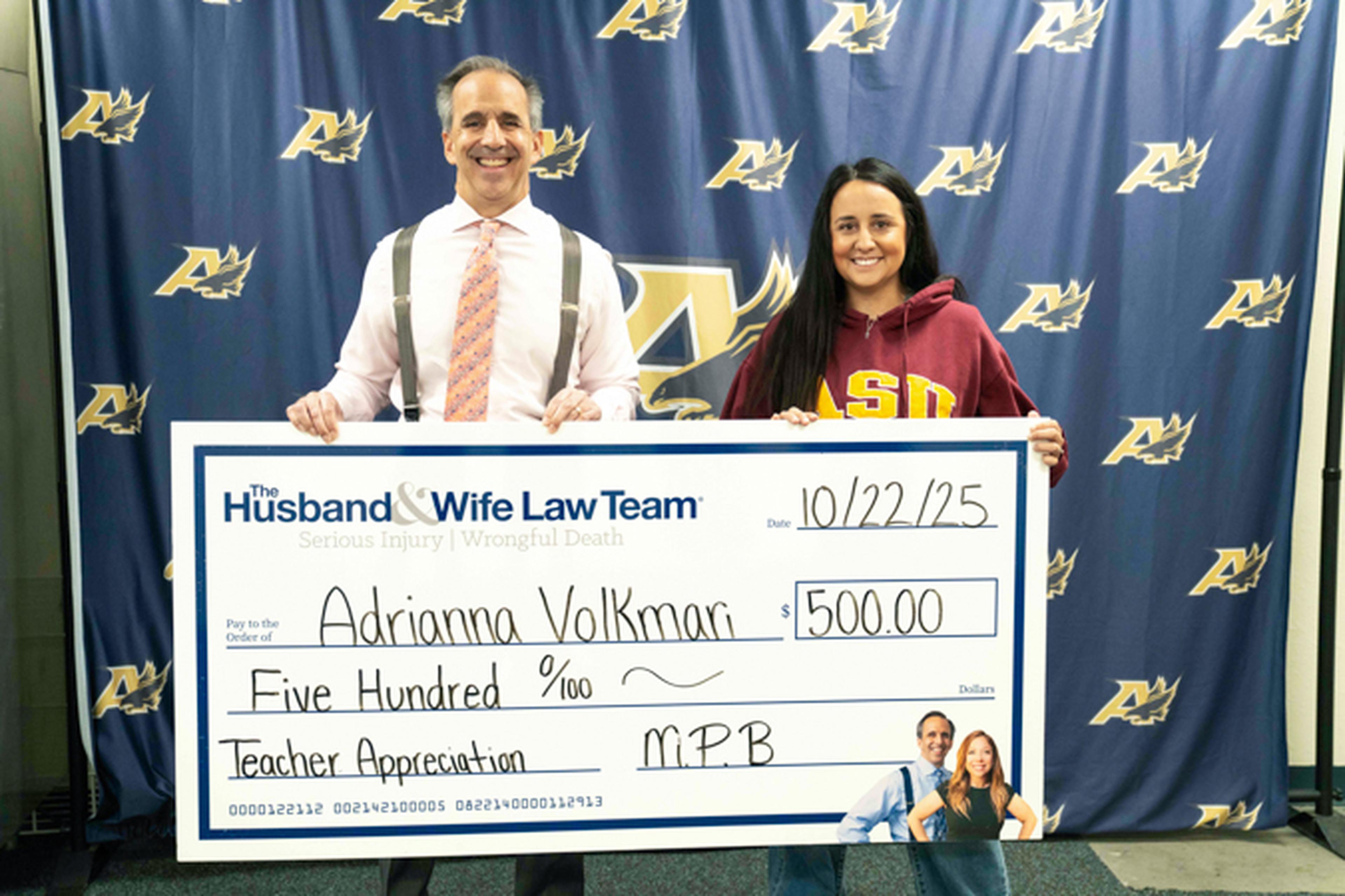 Mark Breyer and the Teacher of the Month hold a large check for $500 in front of a backdrop with logos. The check is for teacher appreciation.