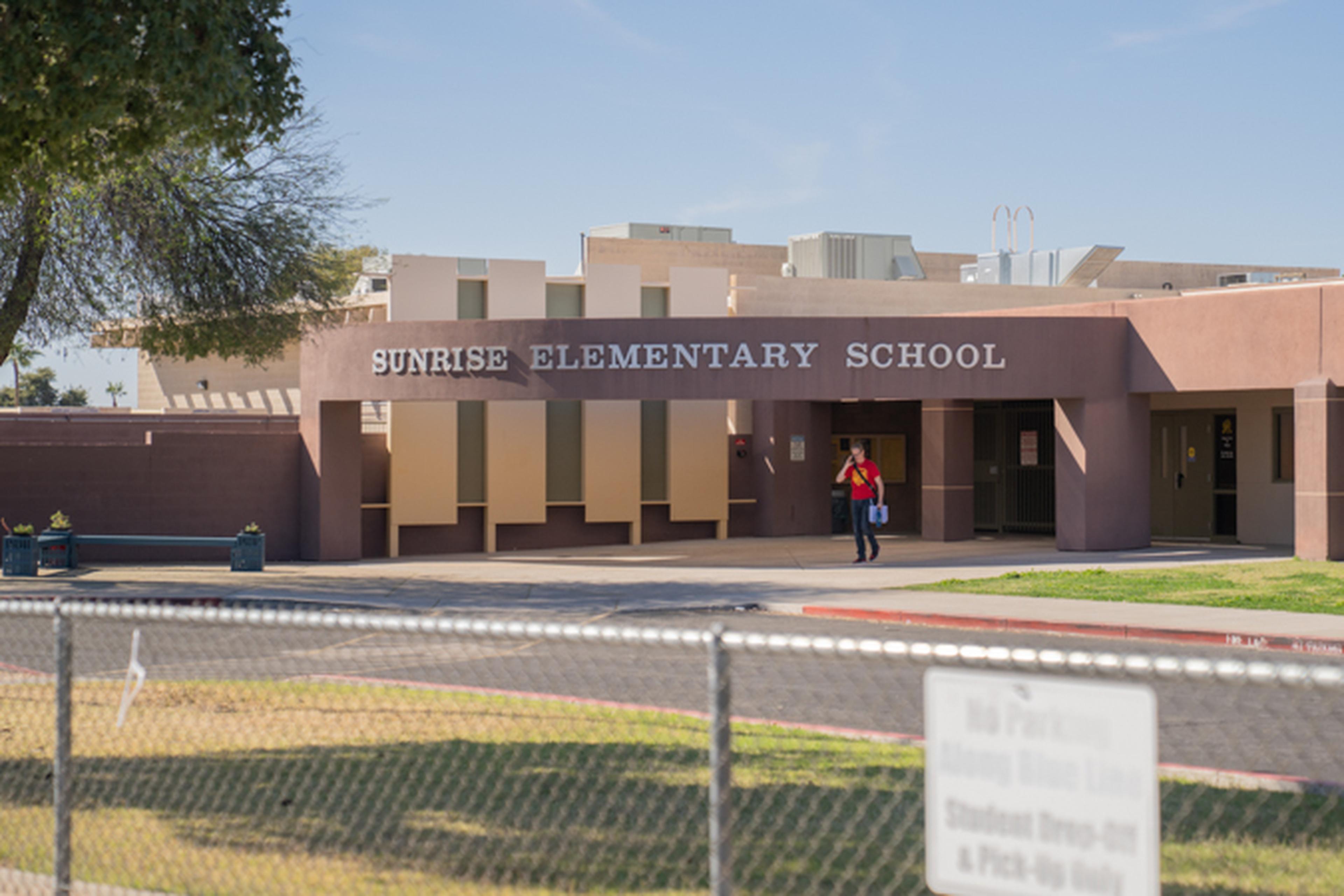 Front view of Sunrise Elementary School building in Phoenix, AZ with a person walking by, a tree on the left, and a chain-link fence in the foreground.