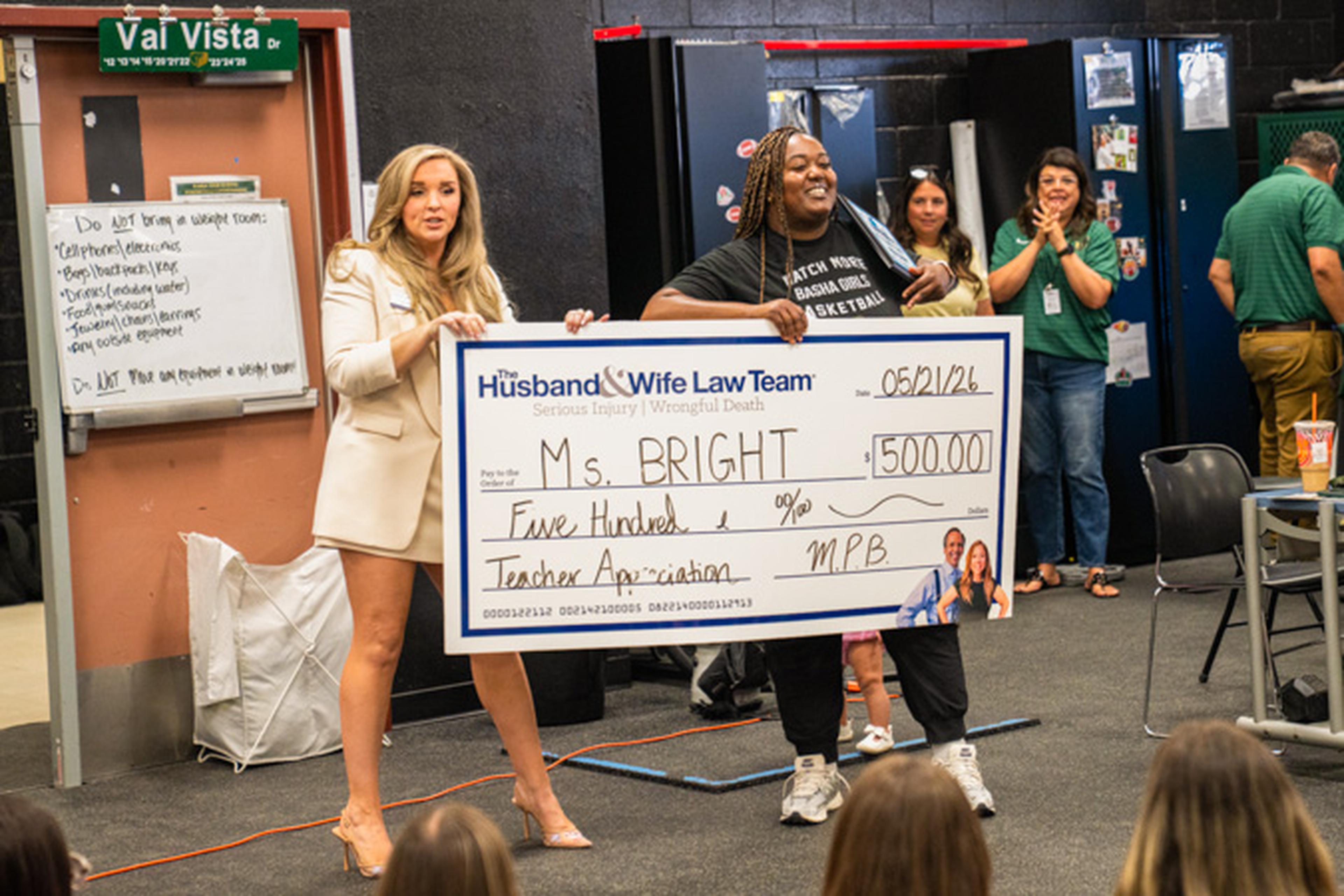 Jen and Coach Bright hold a $500 Teacher Appreciation check at the front of the classroom as students and staff clap proudly in the background.