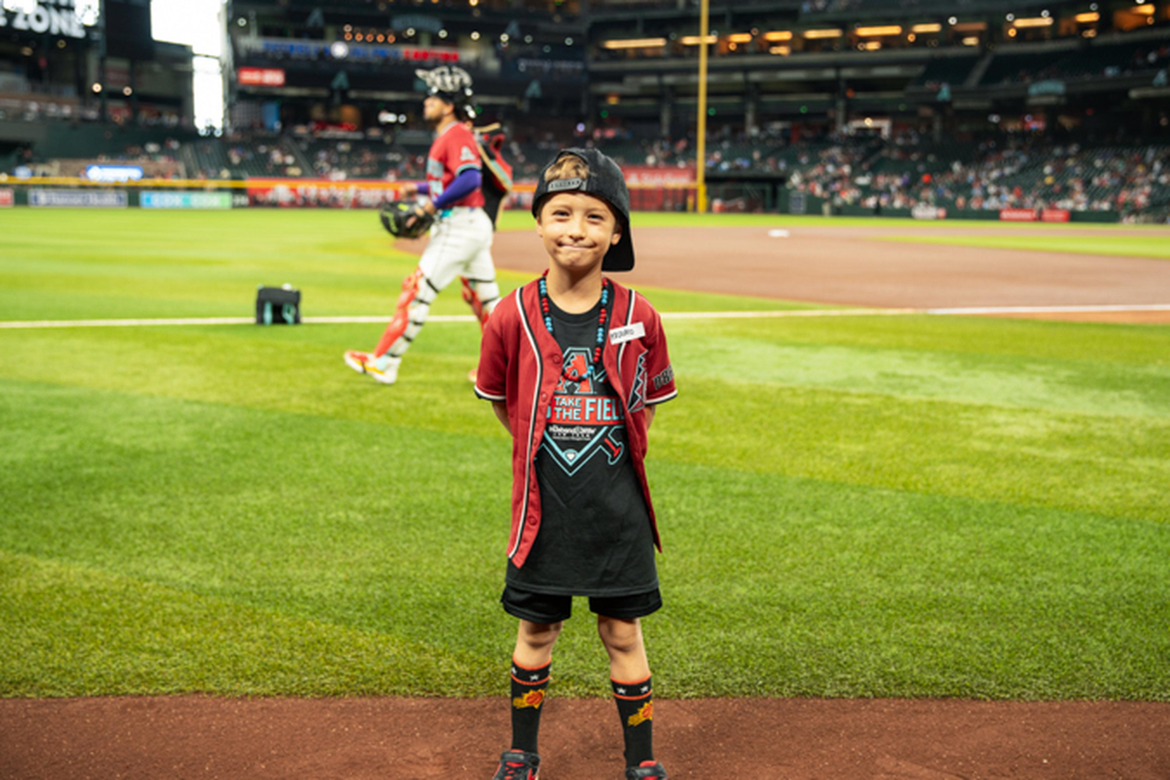 Young boy in a baseball jersey and cap stands on a baseball field, smiling. A player walks in the background. Stadium seats are partially filled.