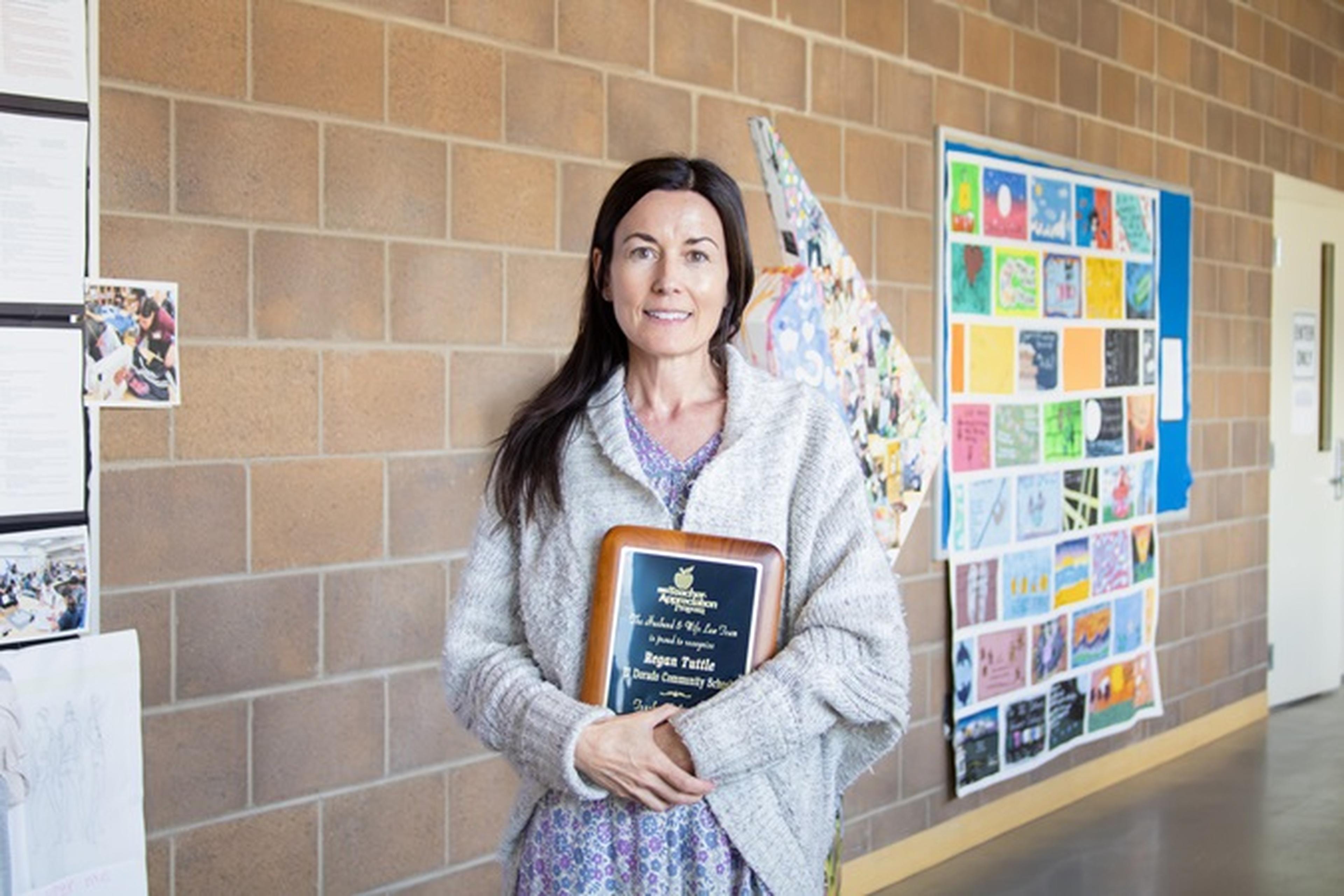 Ms. Tuttle stands smiling with her Teacher Appreciation Award in front of a bulletin board displaying colorful papers and artwork against a brick wall