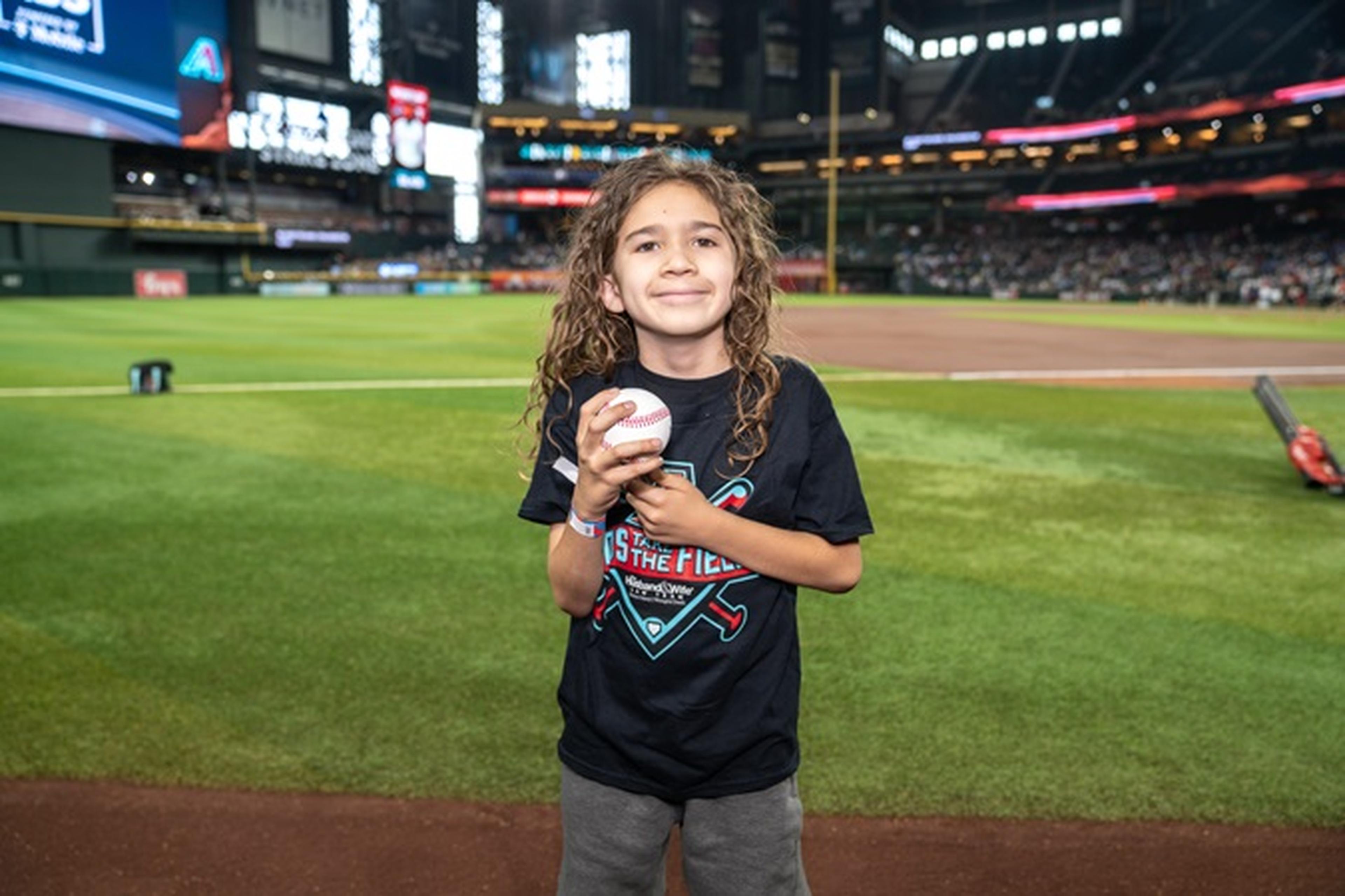 Child with long hair holding a baseball, standing on a field at a stadium, wearing a black shirt and smiling at the camera.