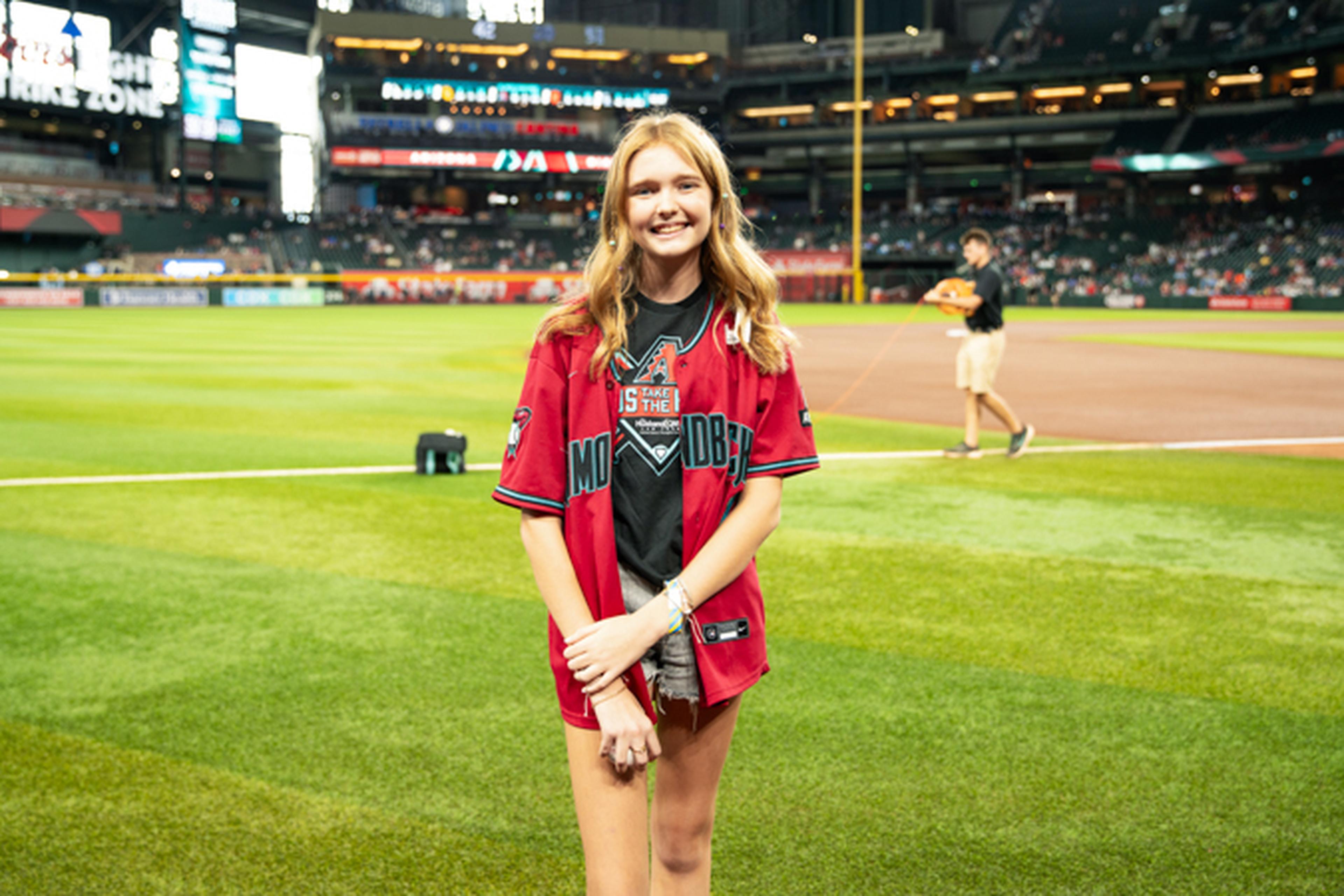 Young woman in a red baseball jersey stands on a baseball field, smiling at the camera. Stadium seats and scoreboard visible in the background.