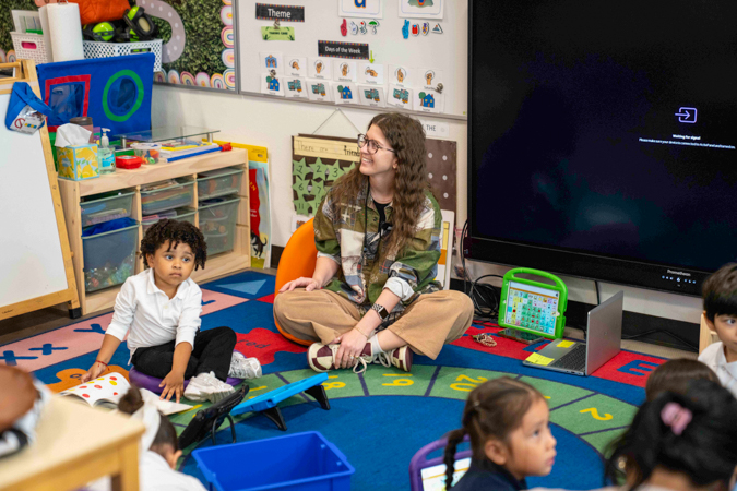 Surprise, Ms. Rachel! We're here to give you an award! Educator sitting on a floor rug with several young kids, smiling