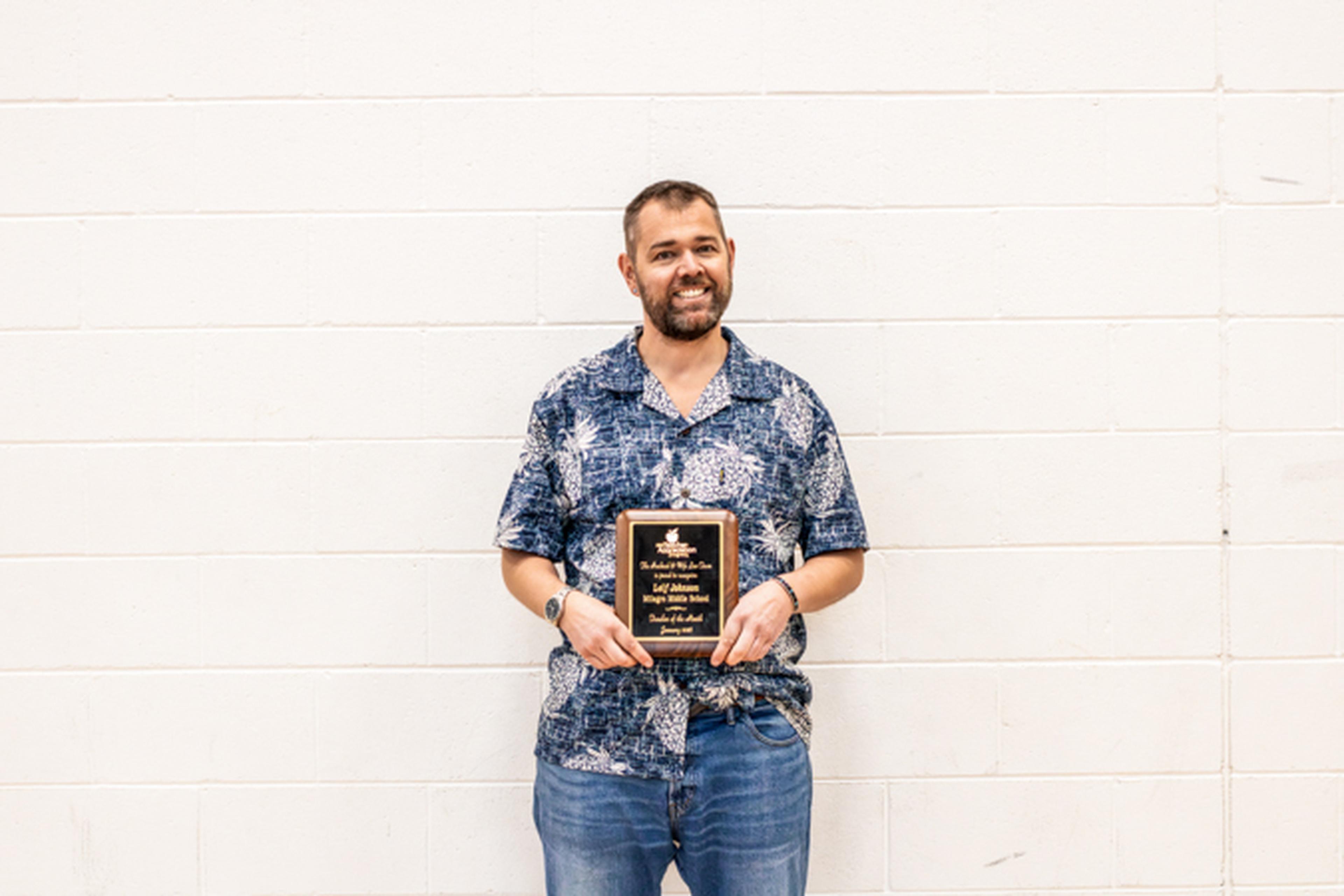 Mr. Johnson holding a plaque, standing in front of a white brick wall.