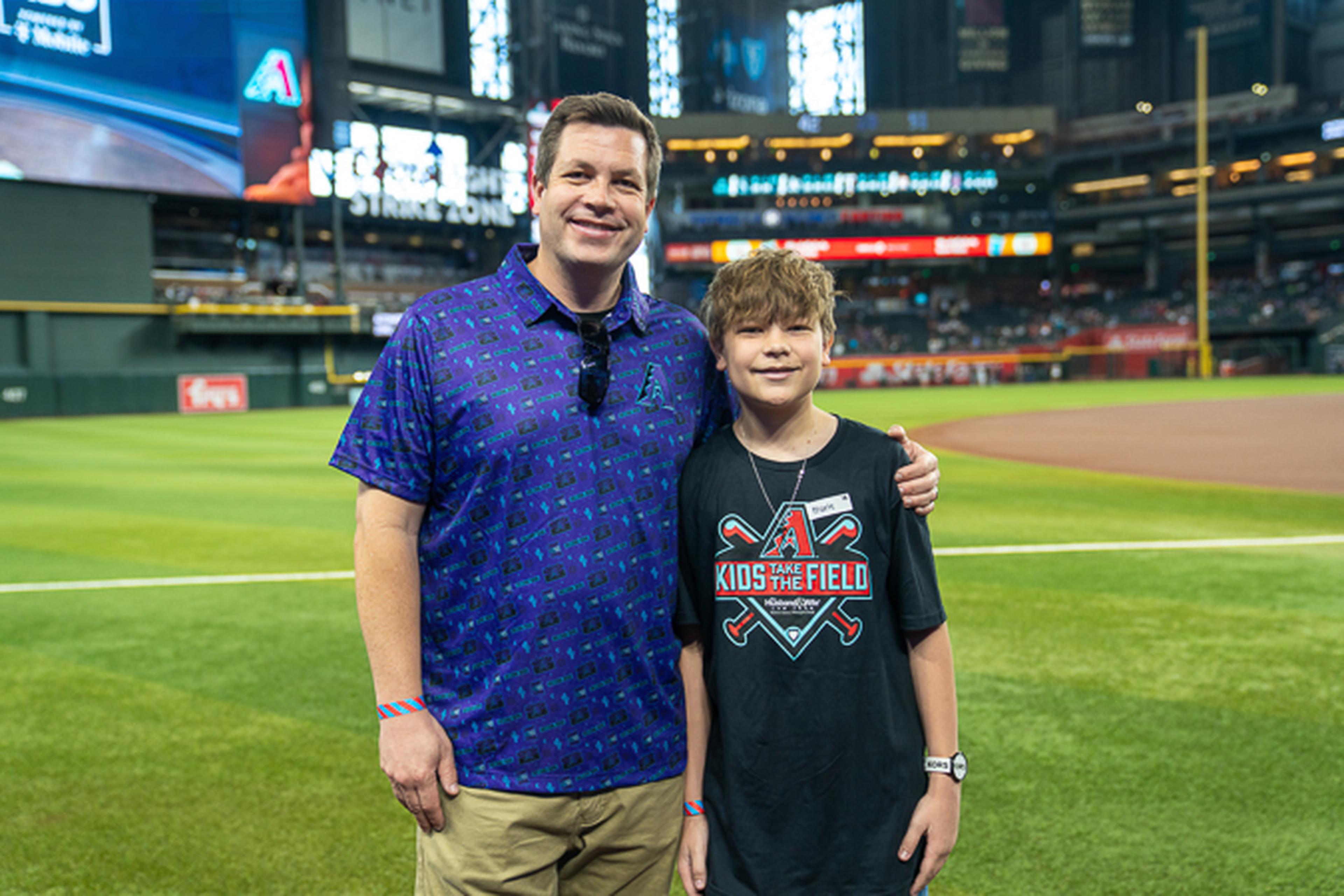 Father and his future MLB player smile on Chase Field with the stadium behind them, the boy proudly wearing his “Kids Take the Field” shirt.