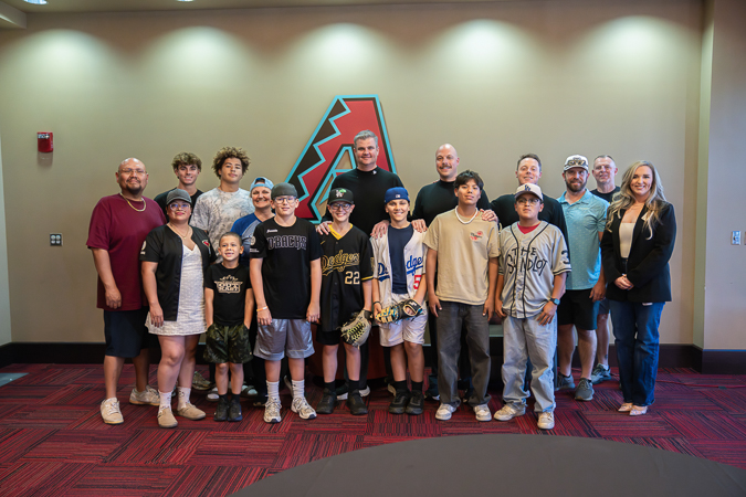 A group of kids and adults, at the UMPS CAREs event, pose smiling indoors against a backdrop with a large red and teal "A" logo.