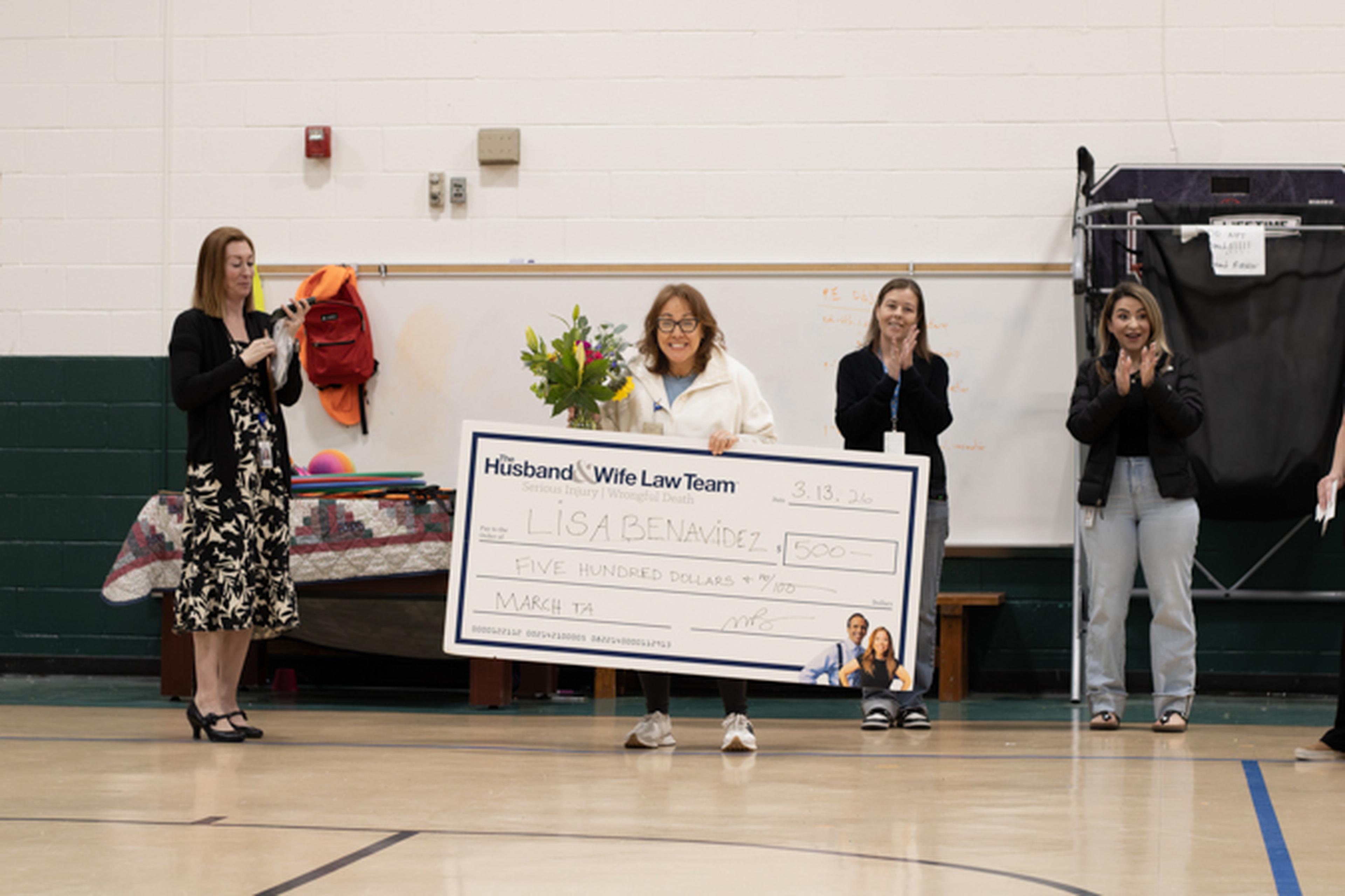 Ms. B holds a large $500 check and a bouquet of flowers from The Husband & Wife Law Team, while three teachers nearby clap and smile in support.