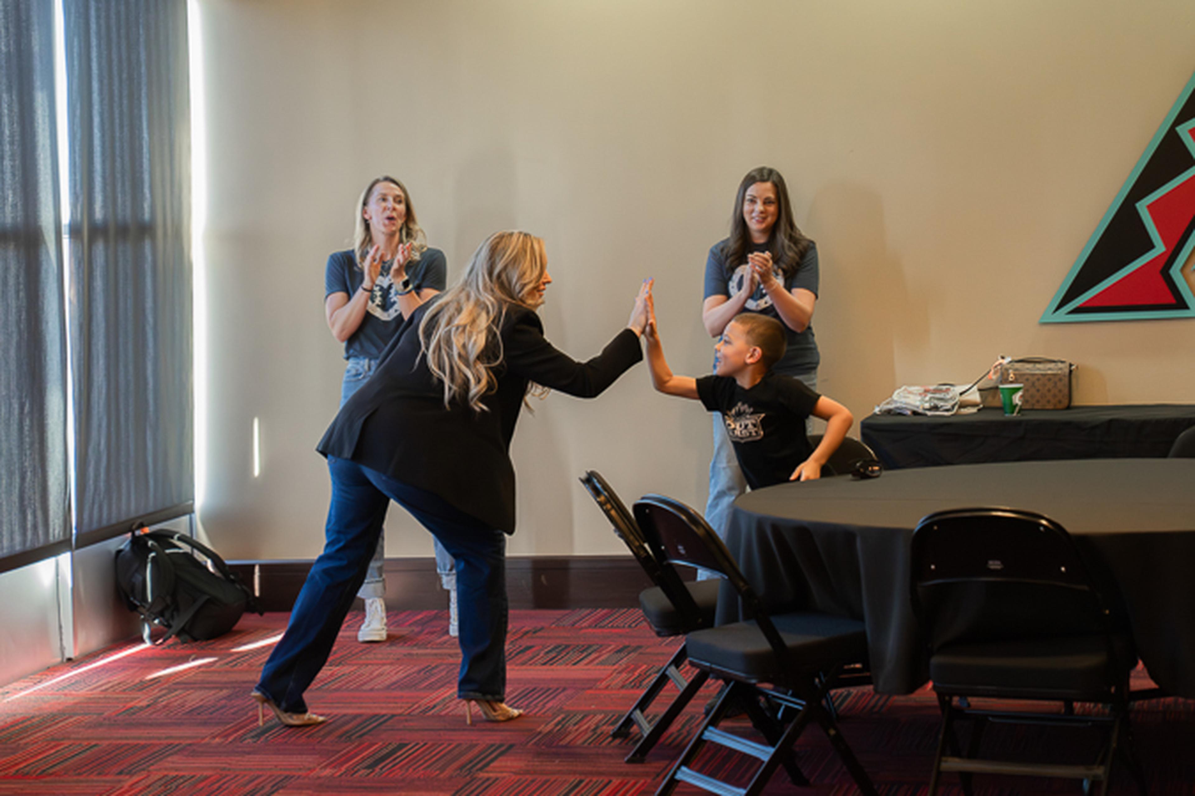 Jen from the Husband & Wife Law Team high‑fives a child at an UMPS CARE event as two women applaud in sunlit background.