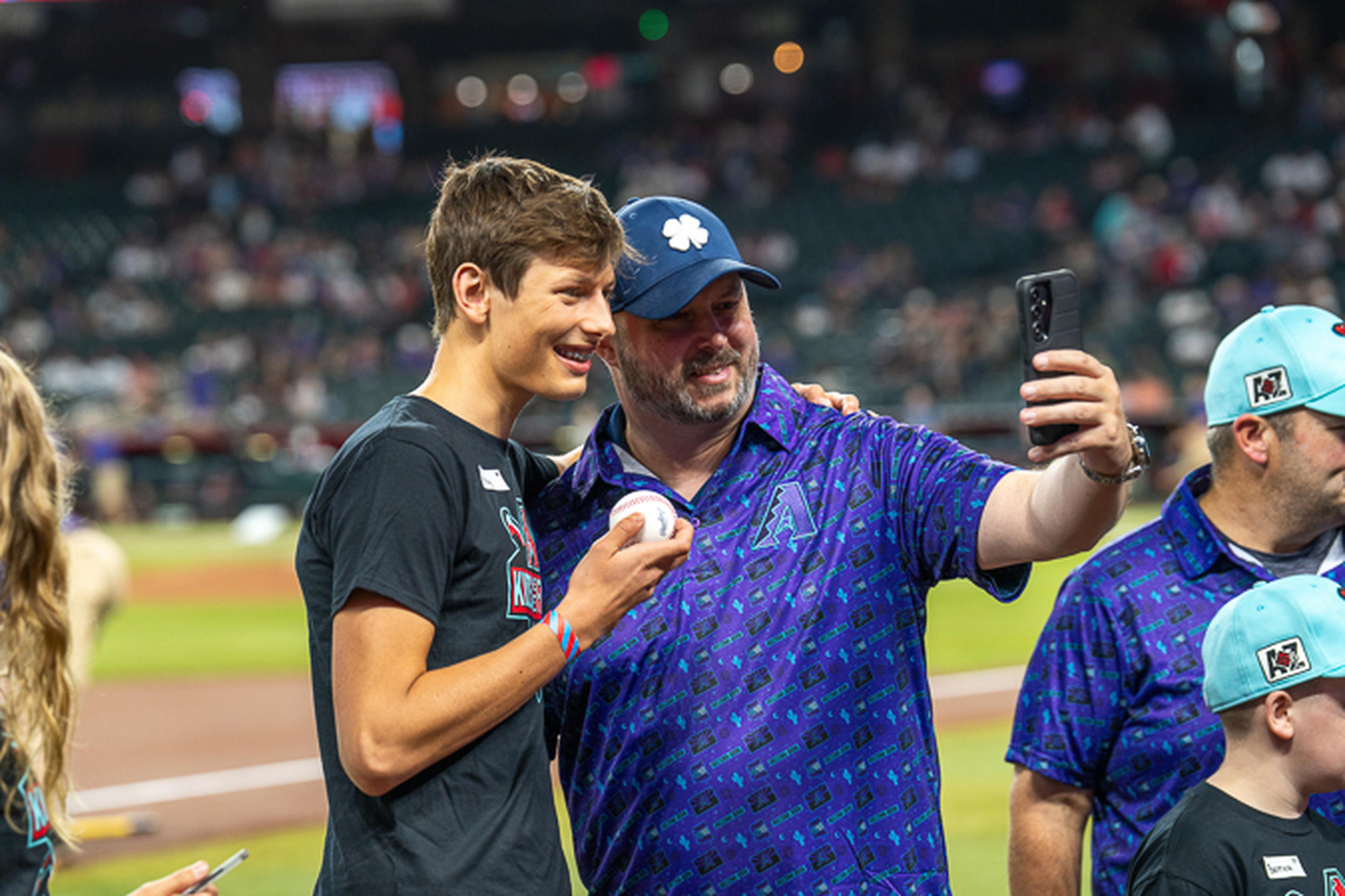 A dad in purple gear and his son in a “Kids on the Field” shirt smile on the baseball field, framed by stadium seating and bat equipment in view.