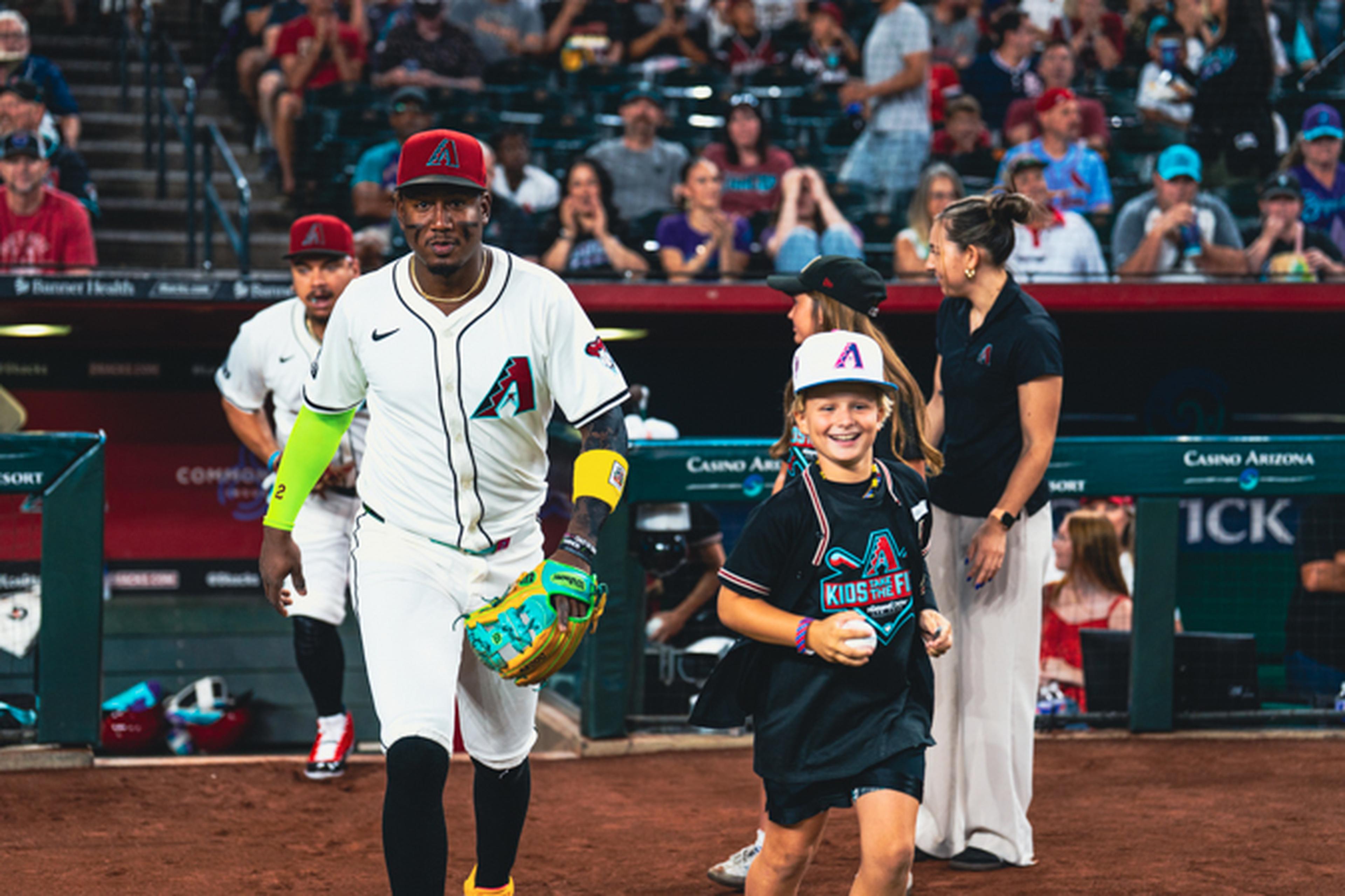 Baseball player and smiling child in team attire walk on field, surrounded by cheering crowd in stadium.