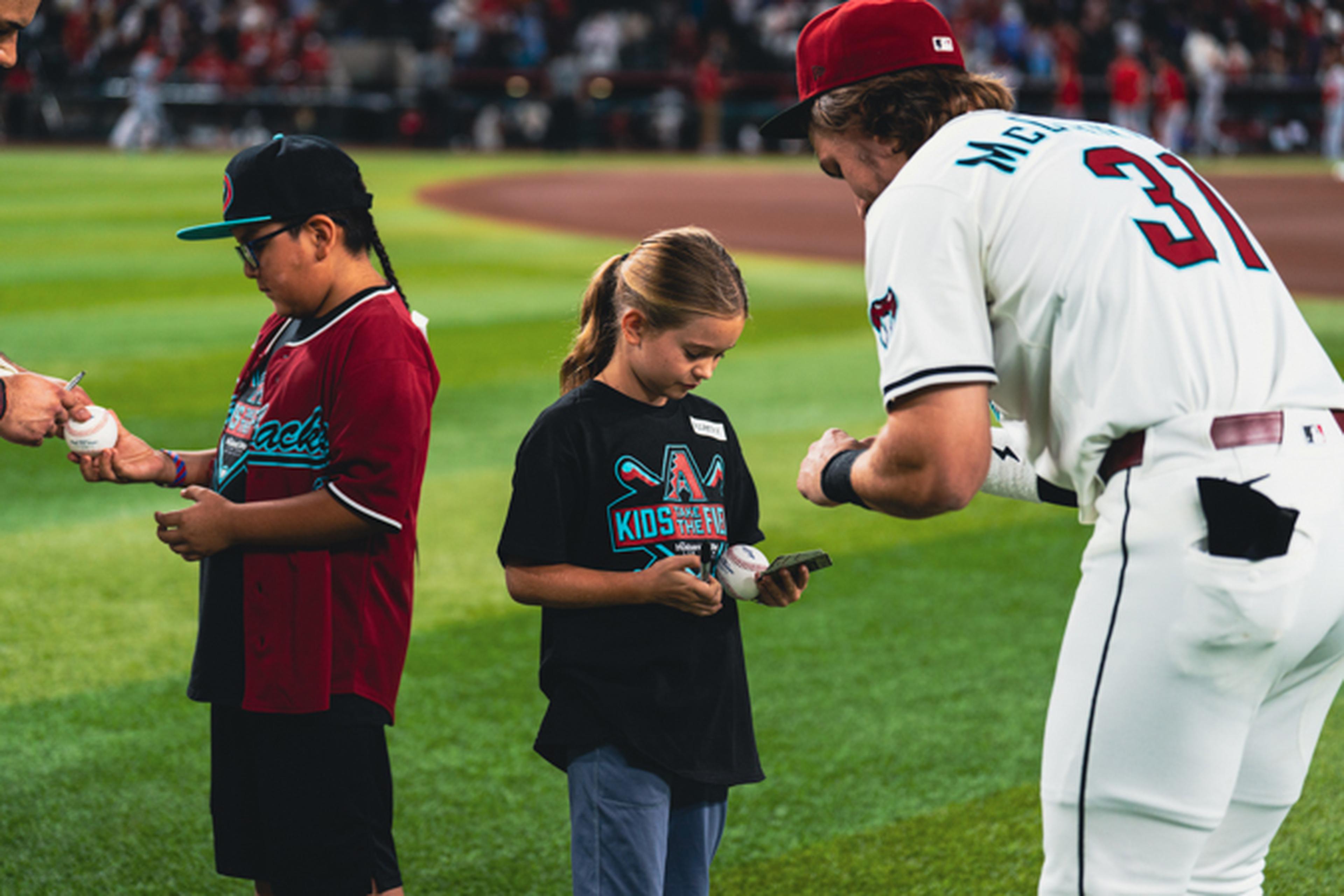 A baseball player signs autographs for two children on a field, with the crowd in the background.