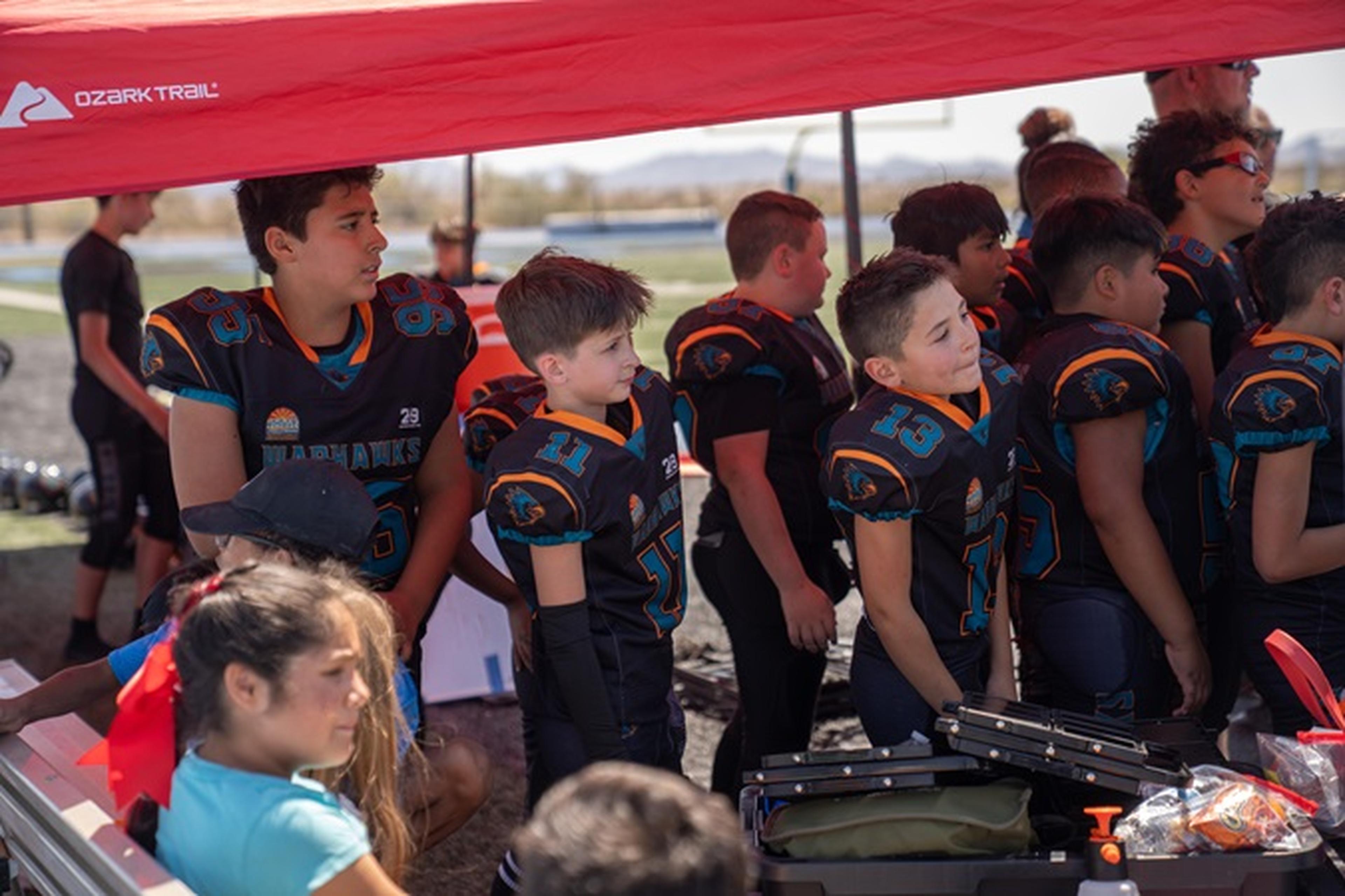 Arizona Youth Football League players in black uniforms gather under a red canopy, focused as they prep for the game with gear laid out on the bench.
