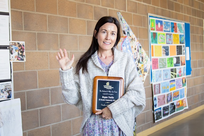 Teacher Appreciation moment — Ms. Tuttle stands proudly with her award before a vibrant bulletin board of student work set against a brick backdrop.