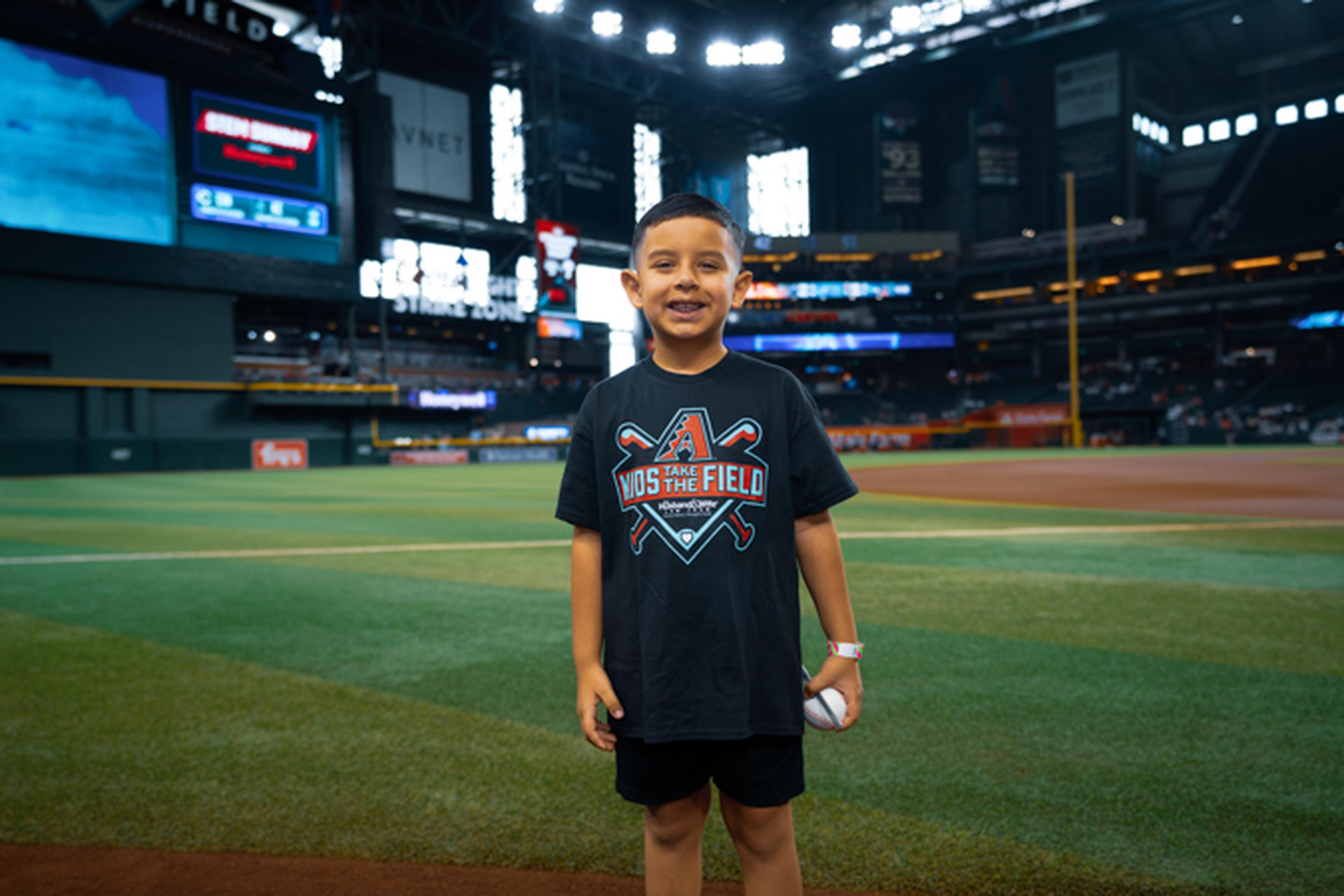 A young boy in a black shirt stands smiling on a baseball field, holding a ball, with a stadium and scoreboard in the background.