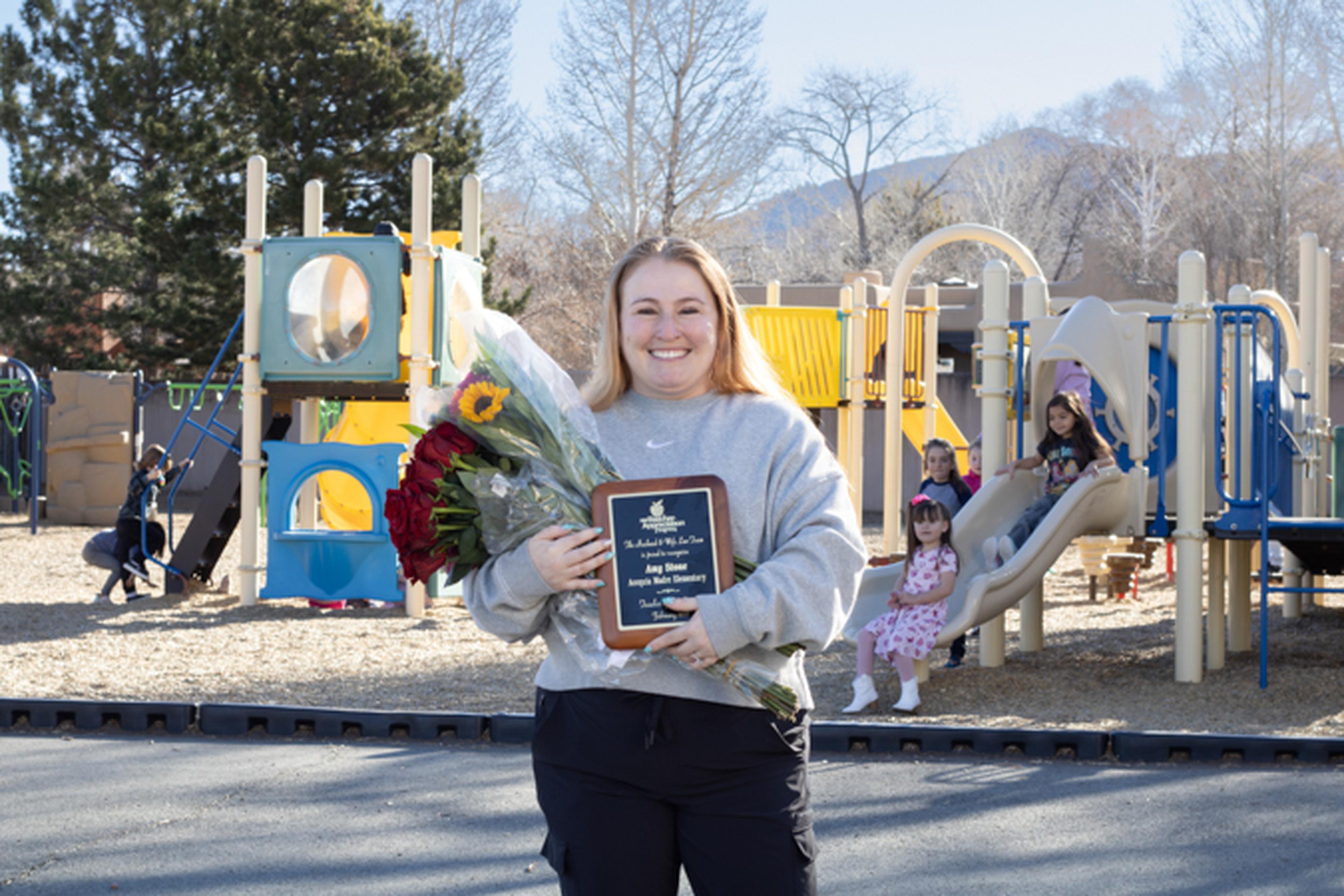 Ms. Amy of Acequia Madre Elementary School holding a plaque and bouquet stands in front of a playground