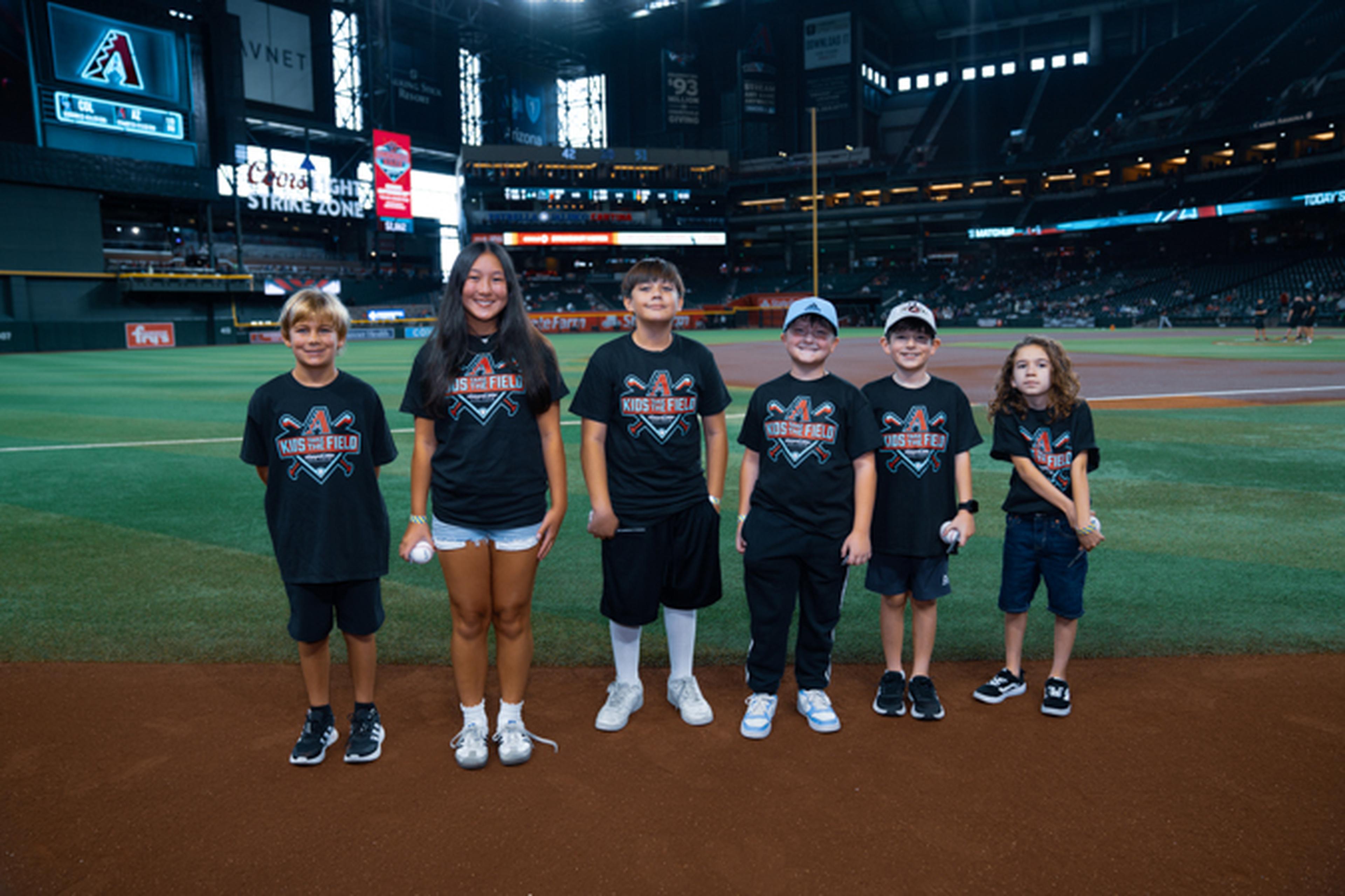 Six children wearing matching baseball-themed shirts stand on a baseball field with a stadium backdrop.