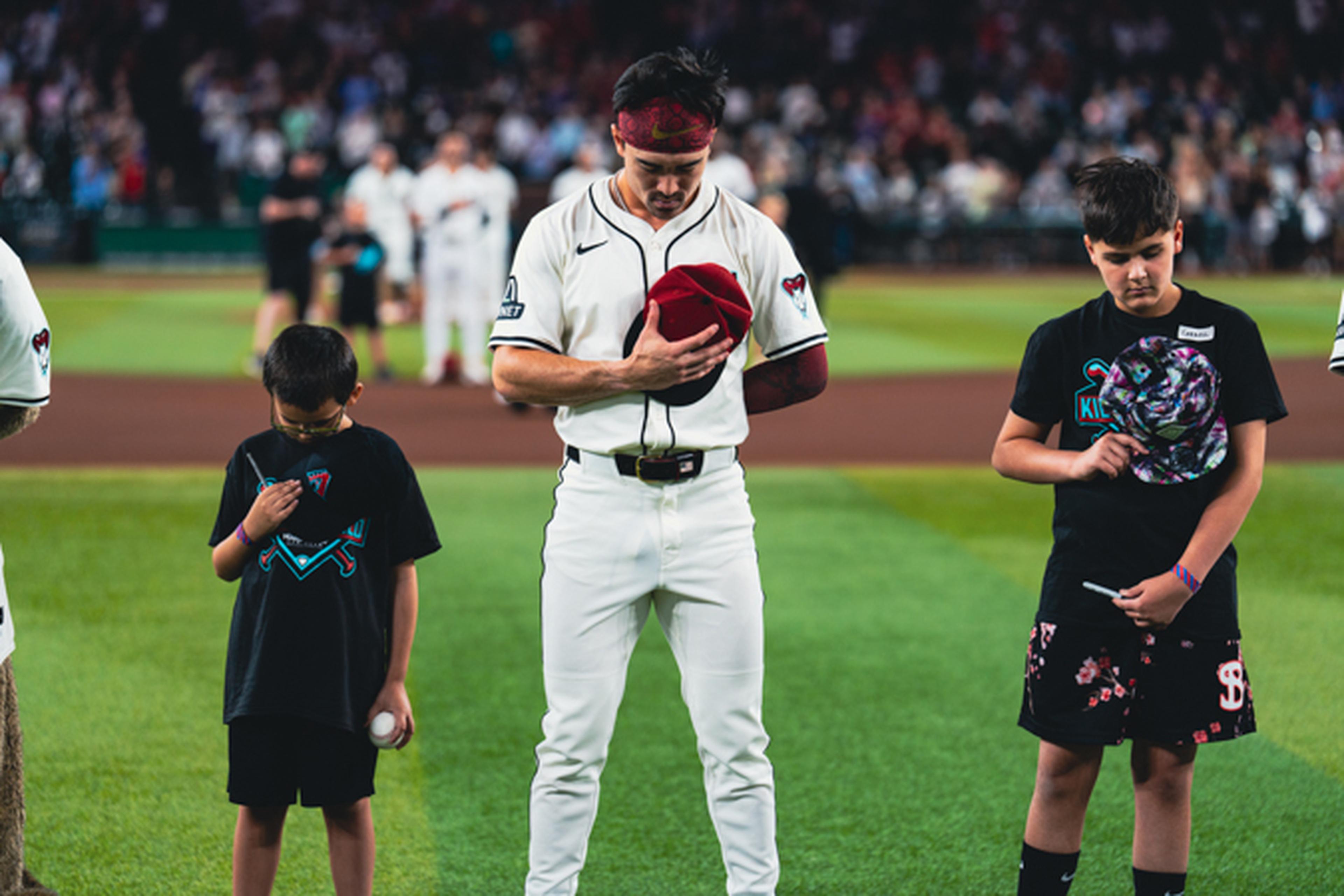 A baseball player and two children stand on a field, heads bowed, holding caps over their hearts, with a crowd in the background.