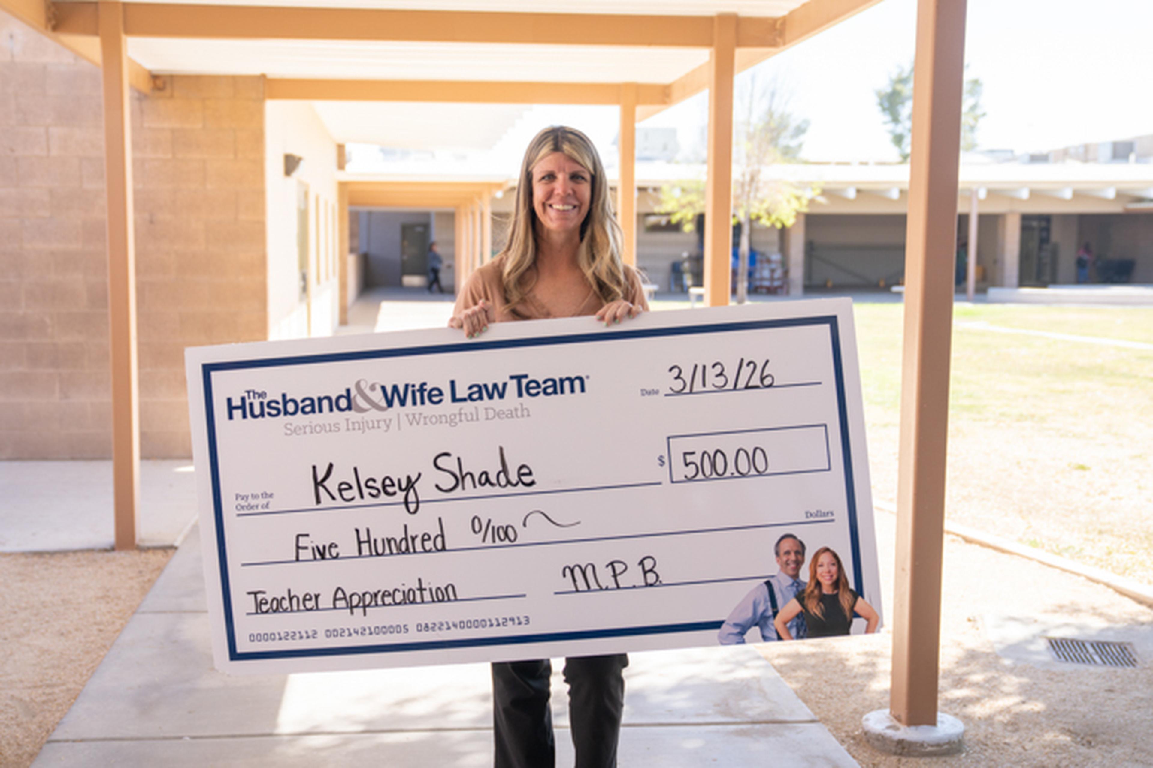 Ms. Shade stands outdoors holding a large check for $500 from "The Husband & Wife Law Team" for her teacher appreciation award.
