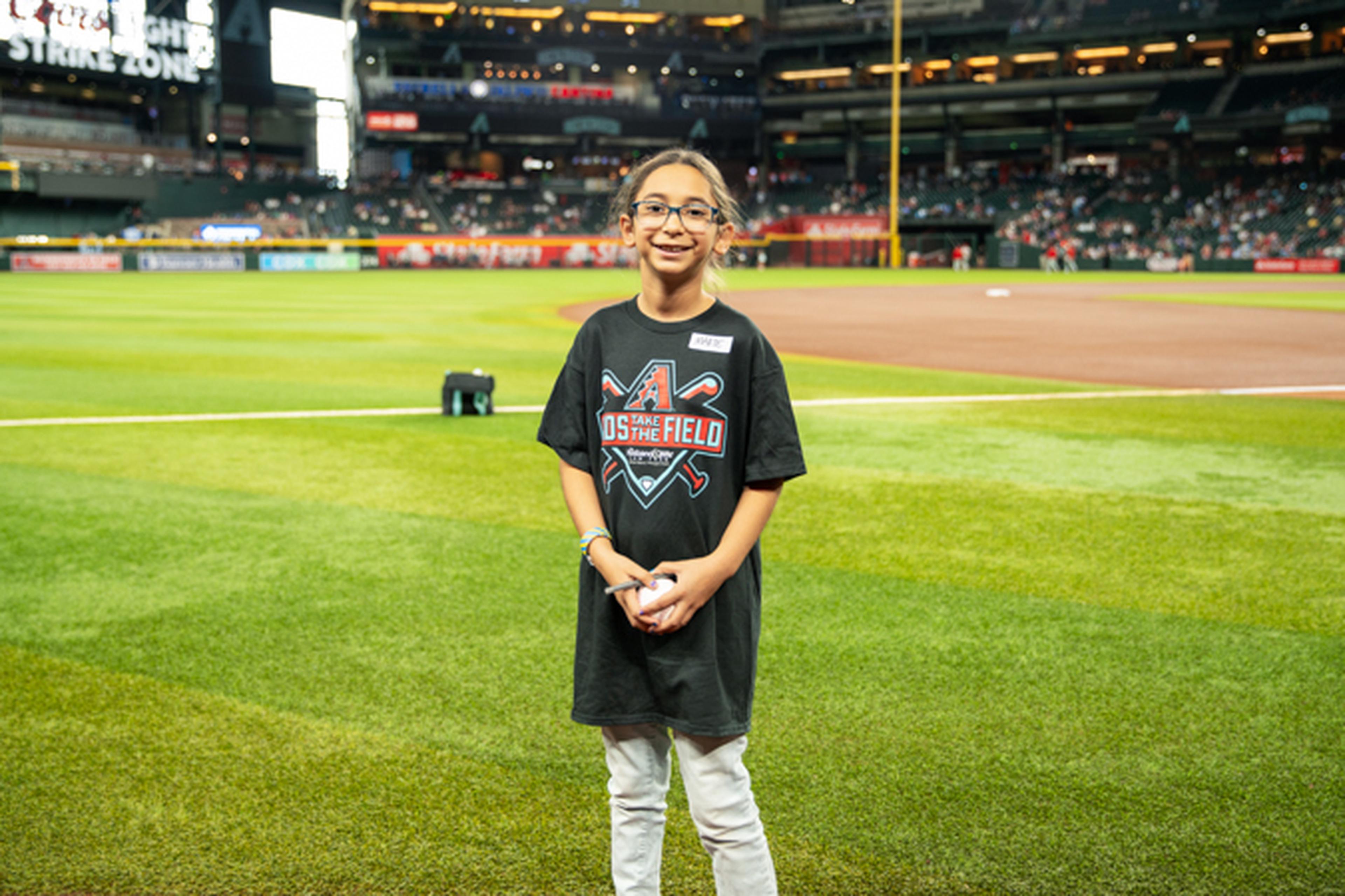 A smiling child stands on a baseball field wearing a black sports-themed T-shirt, with a stadium and crowd in the background.