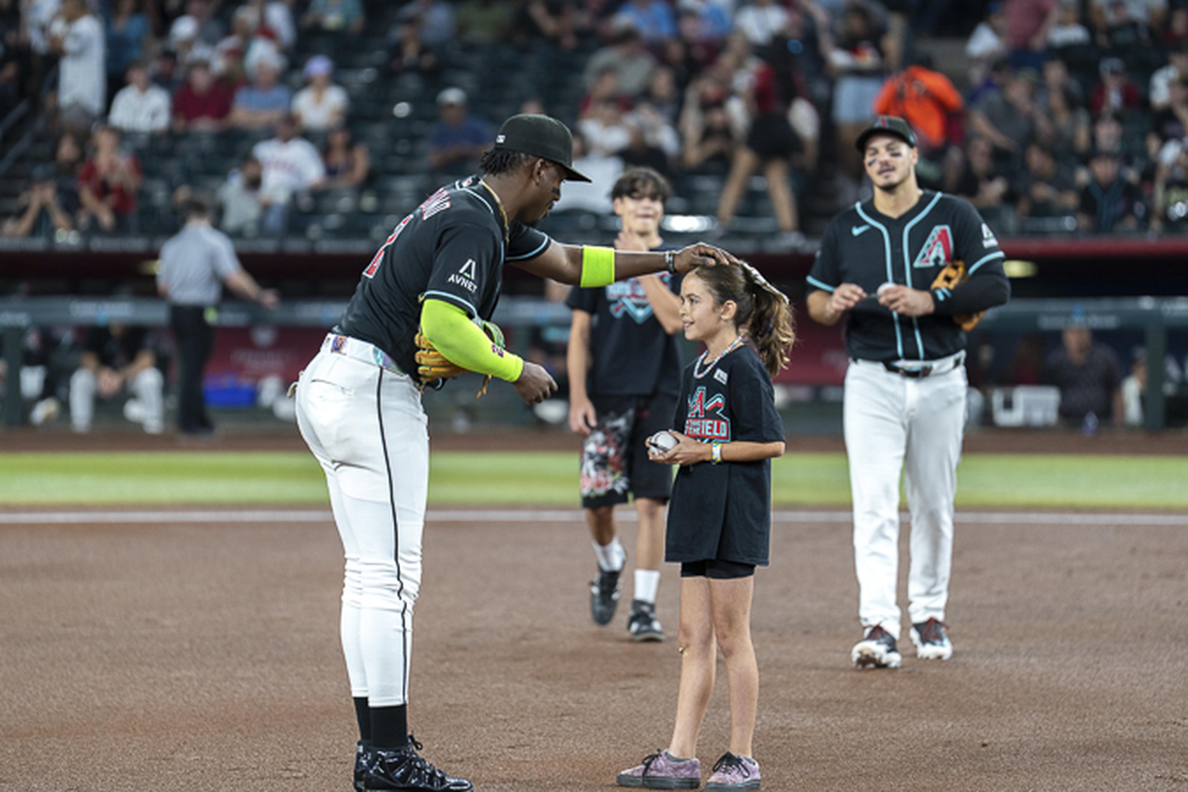 A Diamondbacks player gently pats a young girl’s head on the field as smiling spectators and teammates look on in the background.