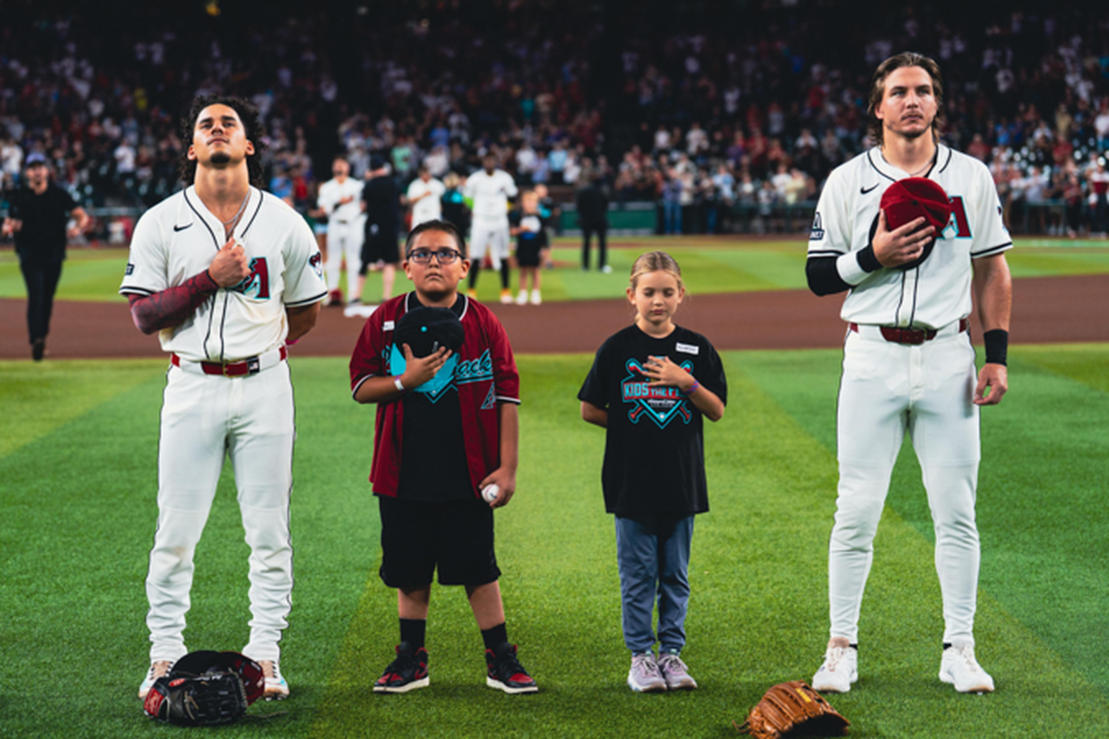 Two baseball players and two children stand on a field with hands over hearts during a national anthem ceremony.
