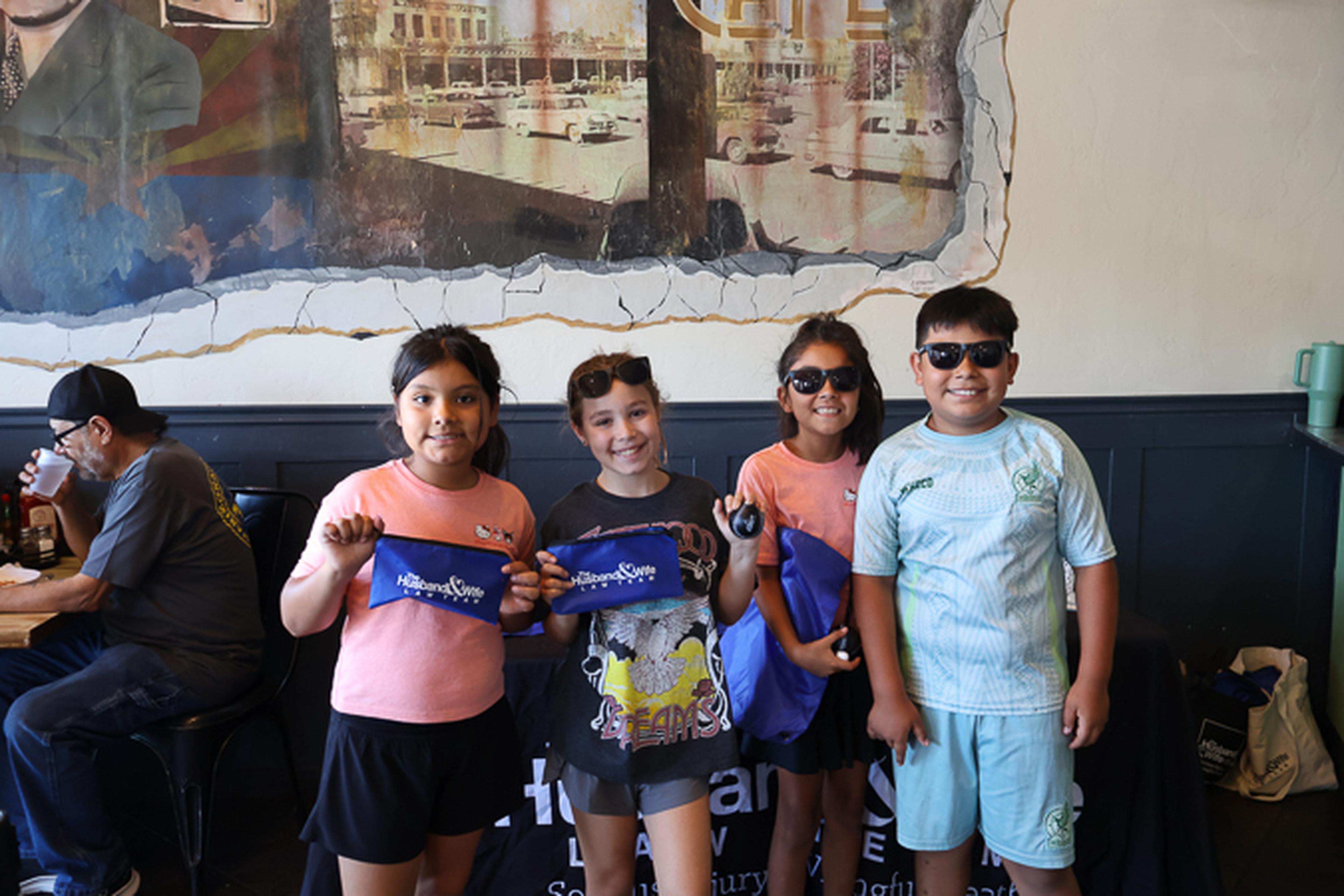 Four young attendees smile while showing off their Husband & Wife Law Team giveaways at Downtown Chandler Cafe & Bakery during a fun community gathering.