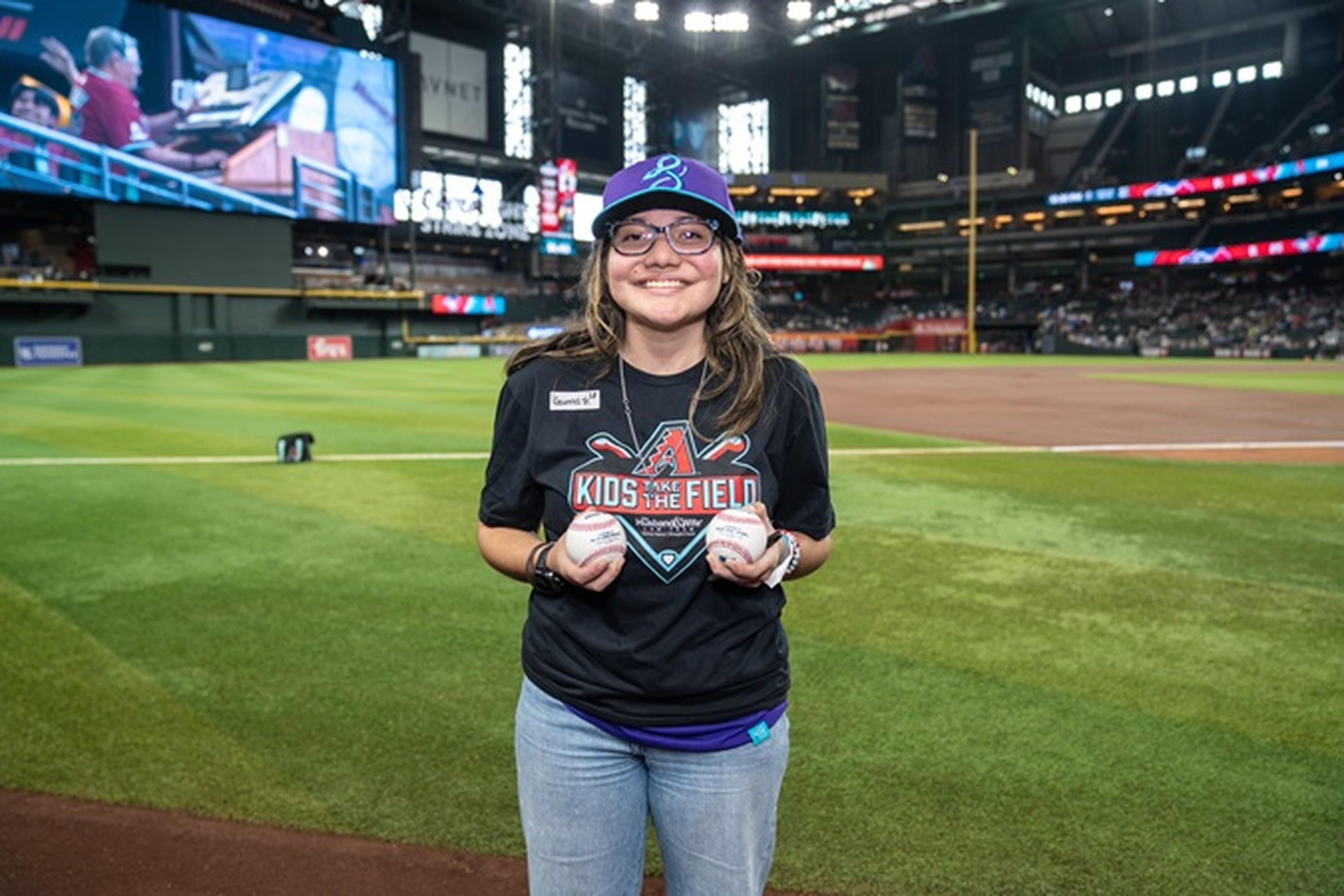 Smiling person wearing a "Kids on the Field" shirt holding baseballs on a baseball field, with a cap and glasses. Stadium in the background.