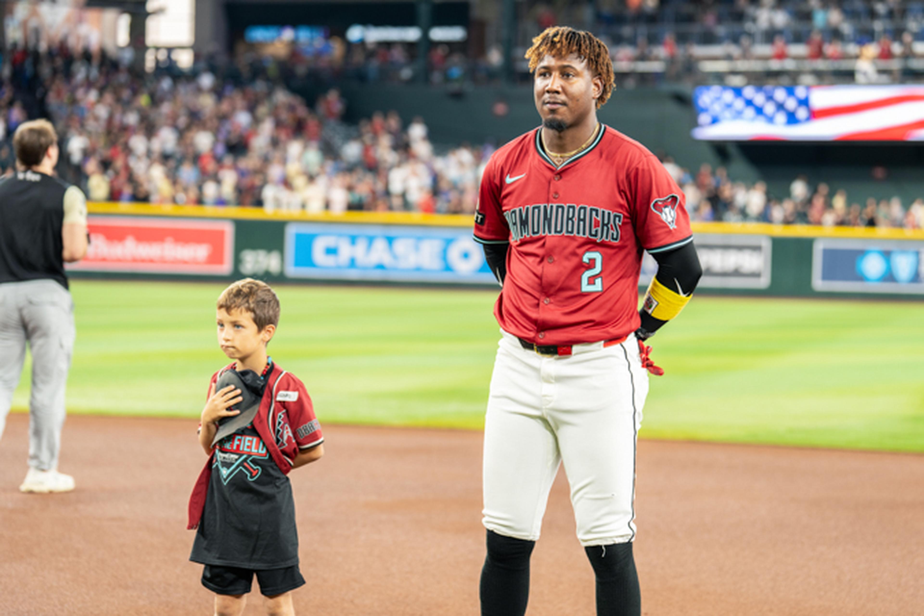Baseball player in a red jersey stands with a young boy on a field during the national anthem, with a crowd and flag in the background.