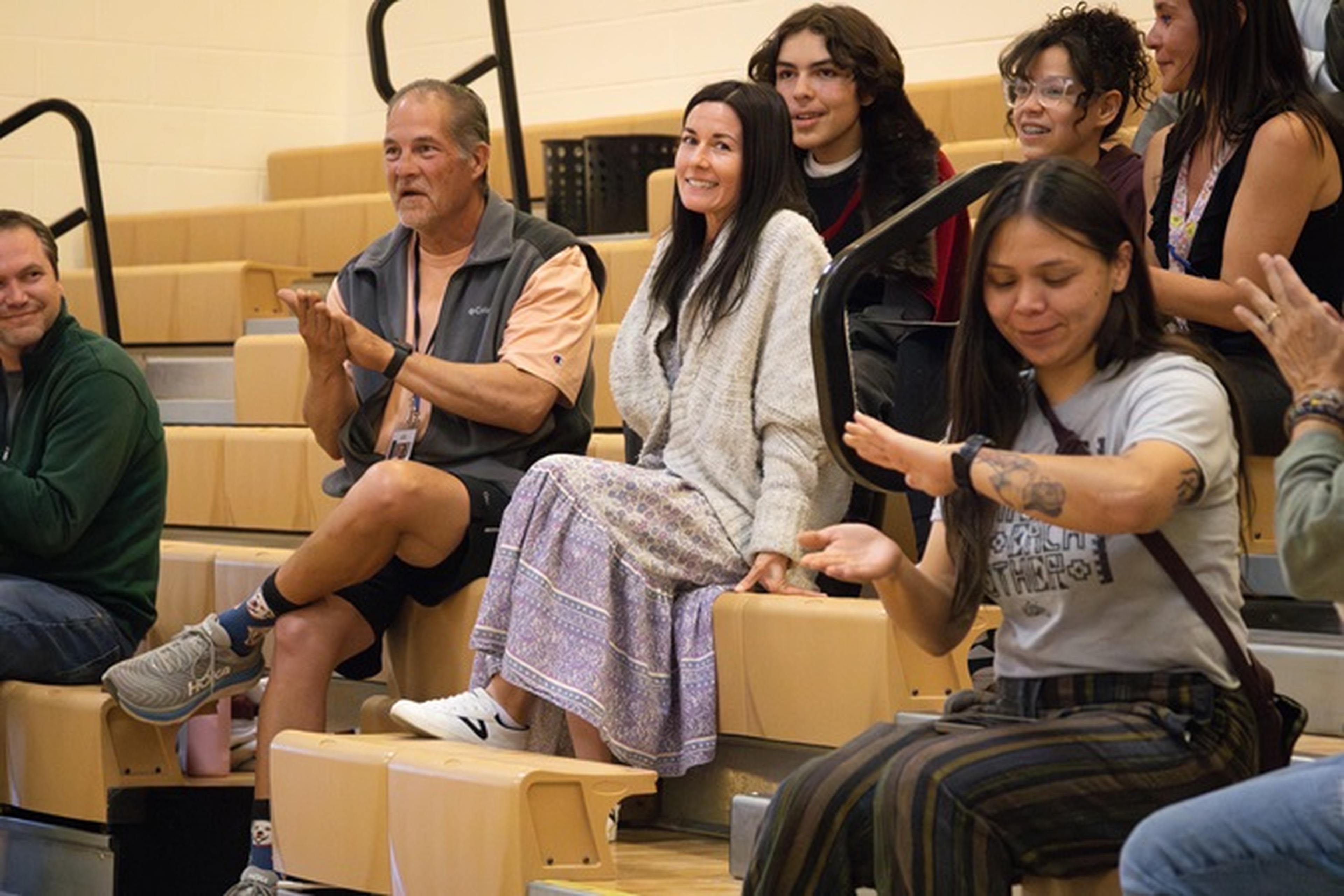Ms. Tuttle and a group of attendees sit together in the bleachers, sharing smiles and applause during the indoor Teacher Appreciation celebration.