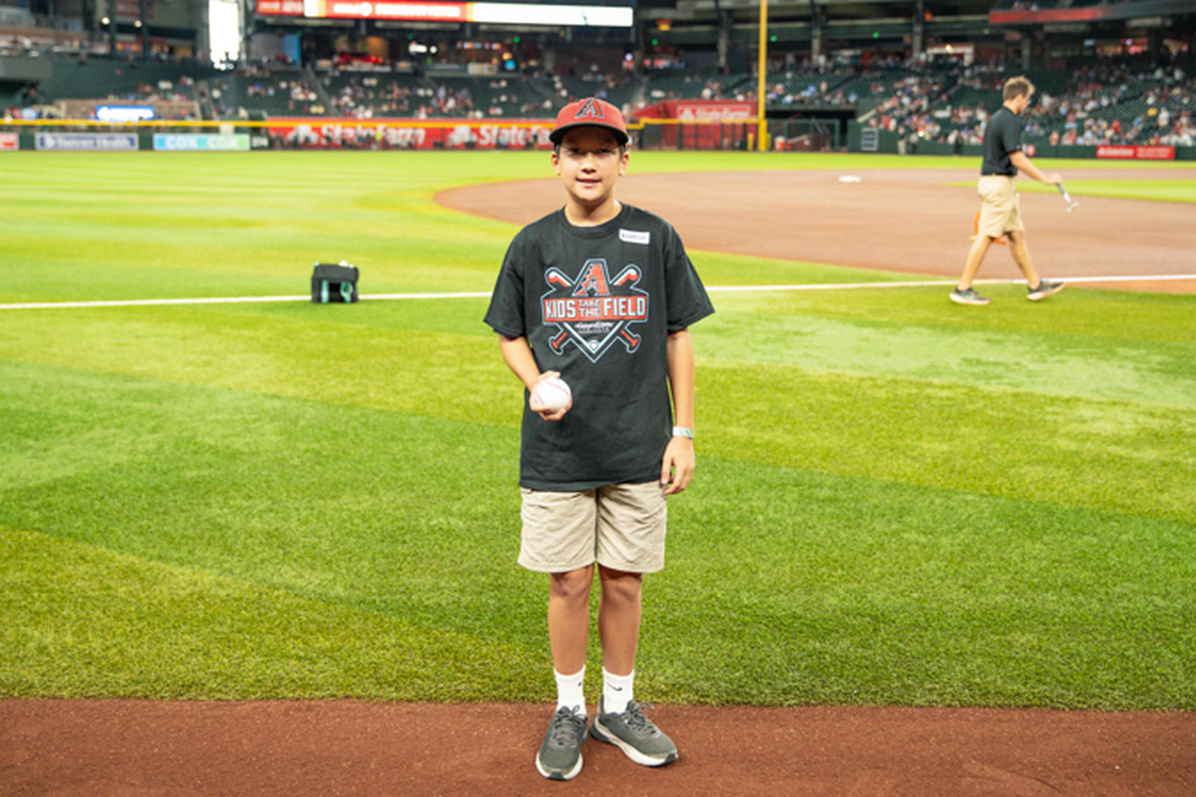 A young boy in a black "Kids on the Field" shirt and shorts holds a baseball, standing on a baseball field near the baseline.