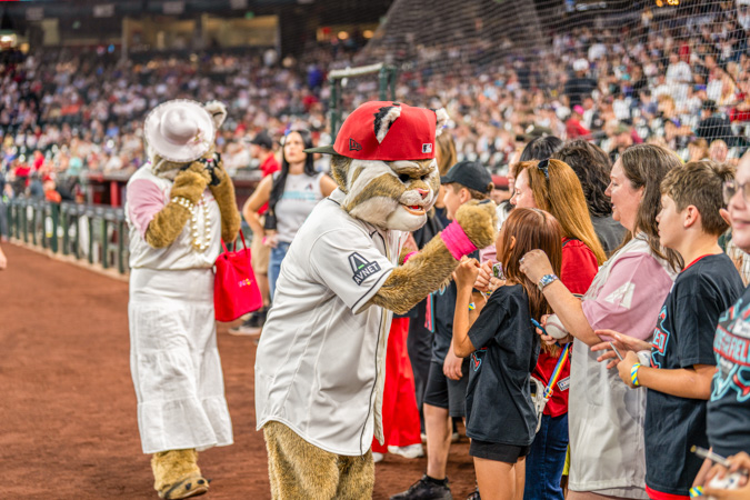 In his bright red cap, Baxter hypes up the crowd near the field while his mom, dressed in white, watches proudly from the background. Baxter the Bobcat entertains excited fans by the diamond, with his mom standing behind him in white, taking in the fun.