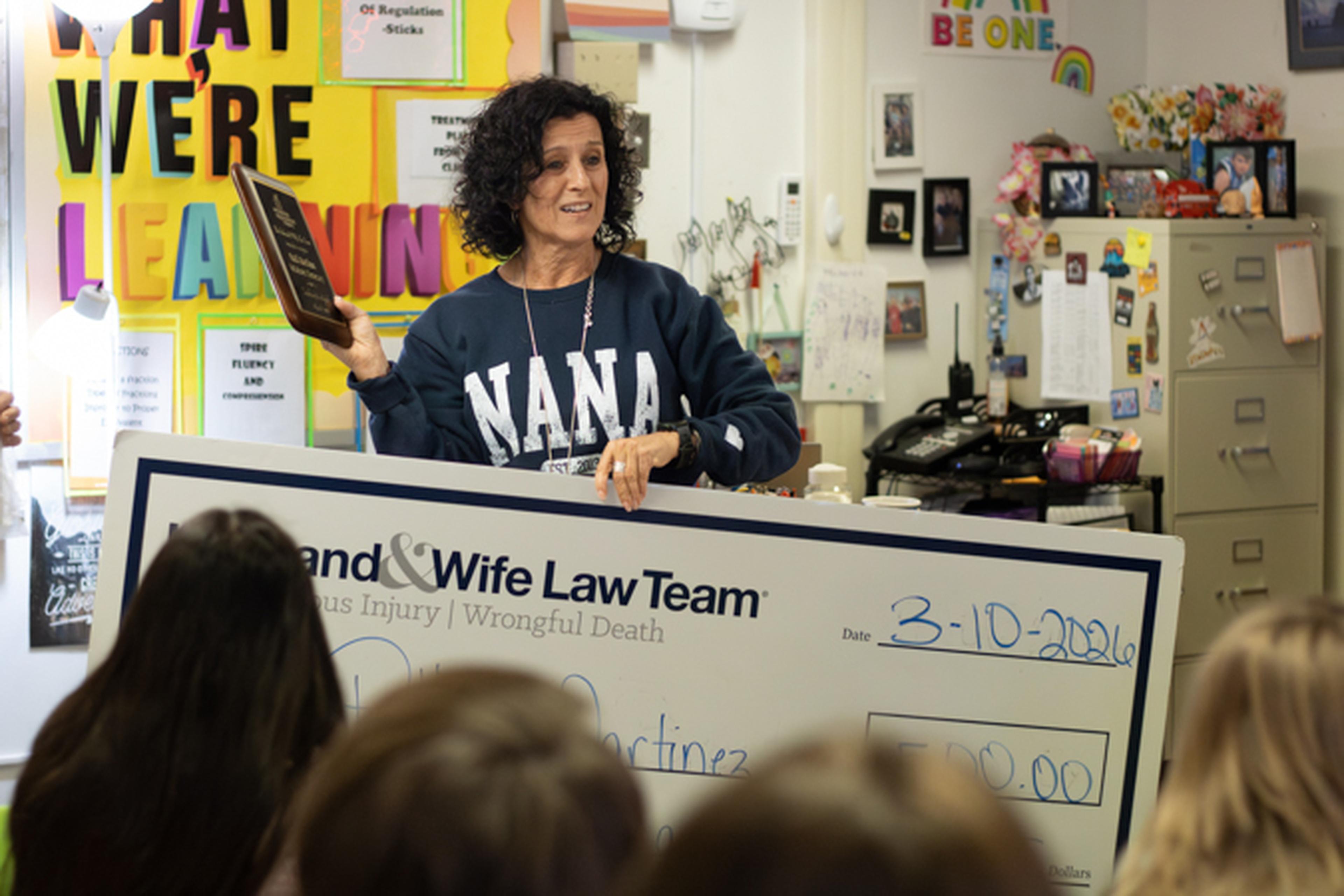 Ms. Martinez of Albuquerque’s Bellehaven Elementary School holds a plaque and a large check for $500 in a decorated room with students facing her.