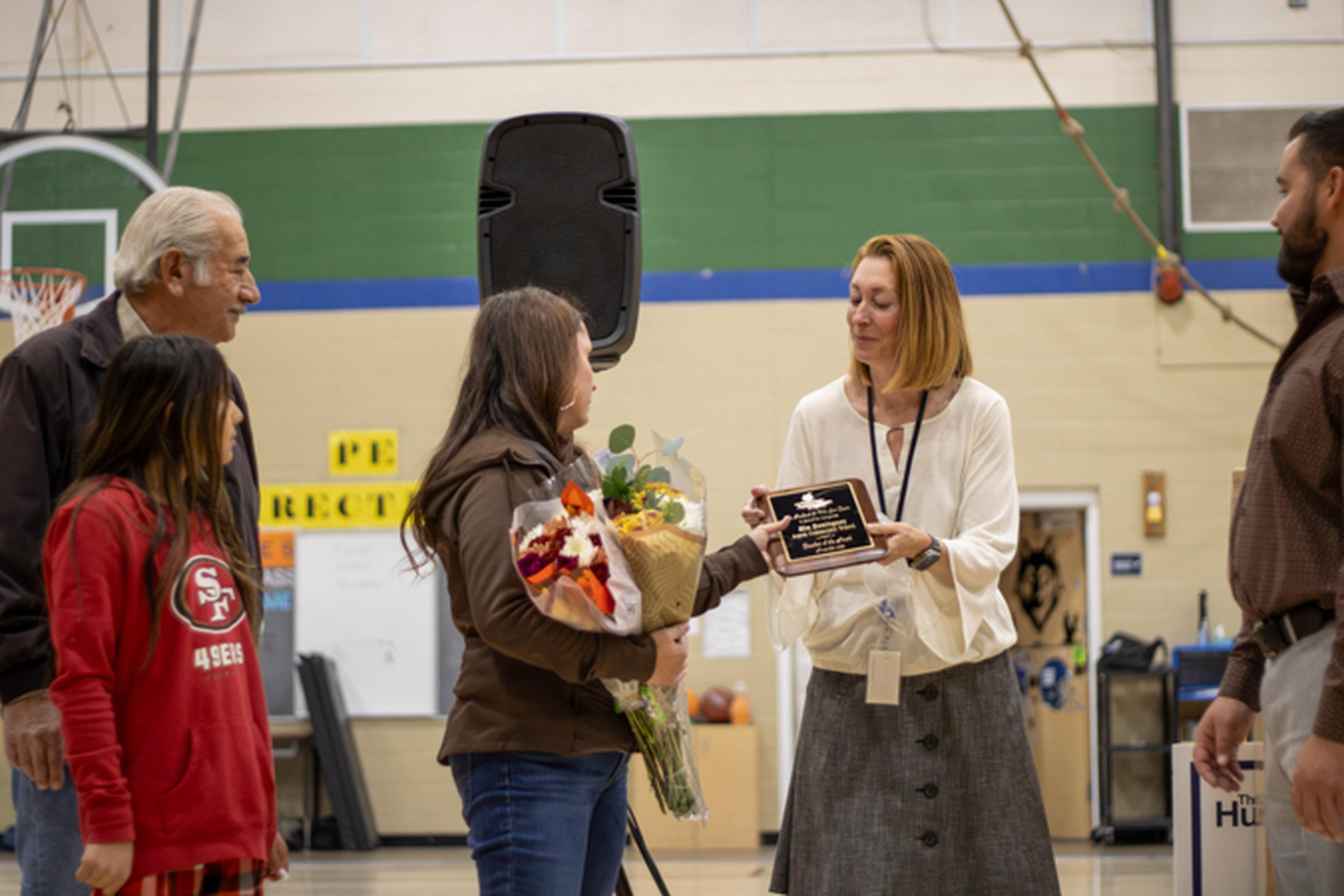 School staff member presents a plaque to a student holding a bouquet in a gym as family watches.