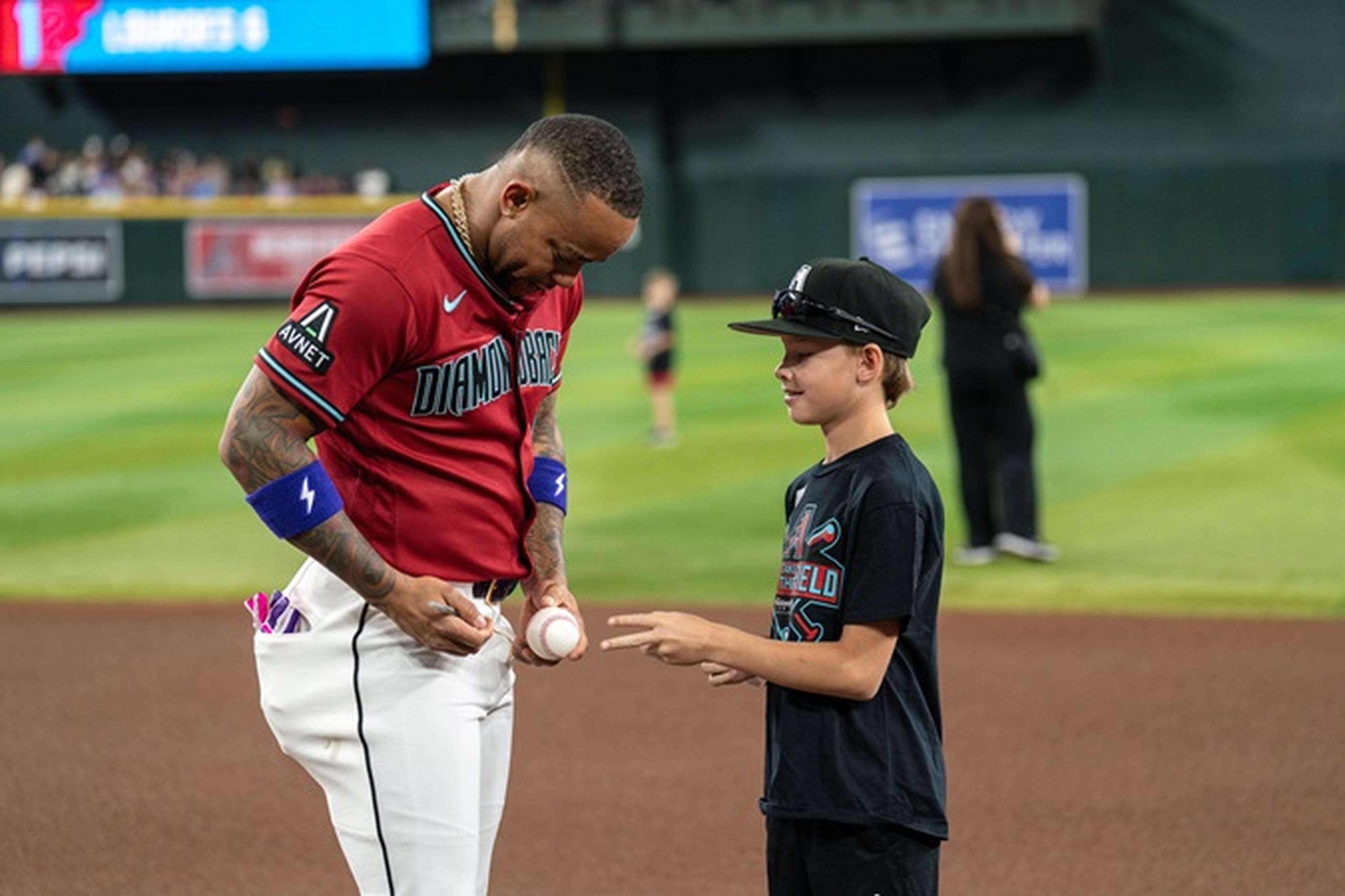 Baseball player in a red jersey signs a baseball for a young fan at a stadium, with green grass and other people in the background.