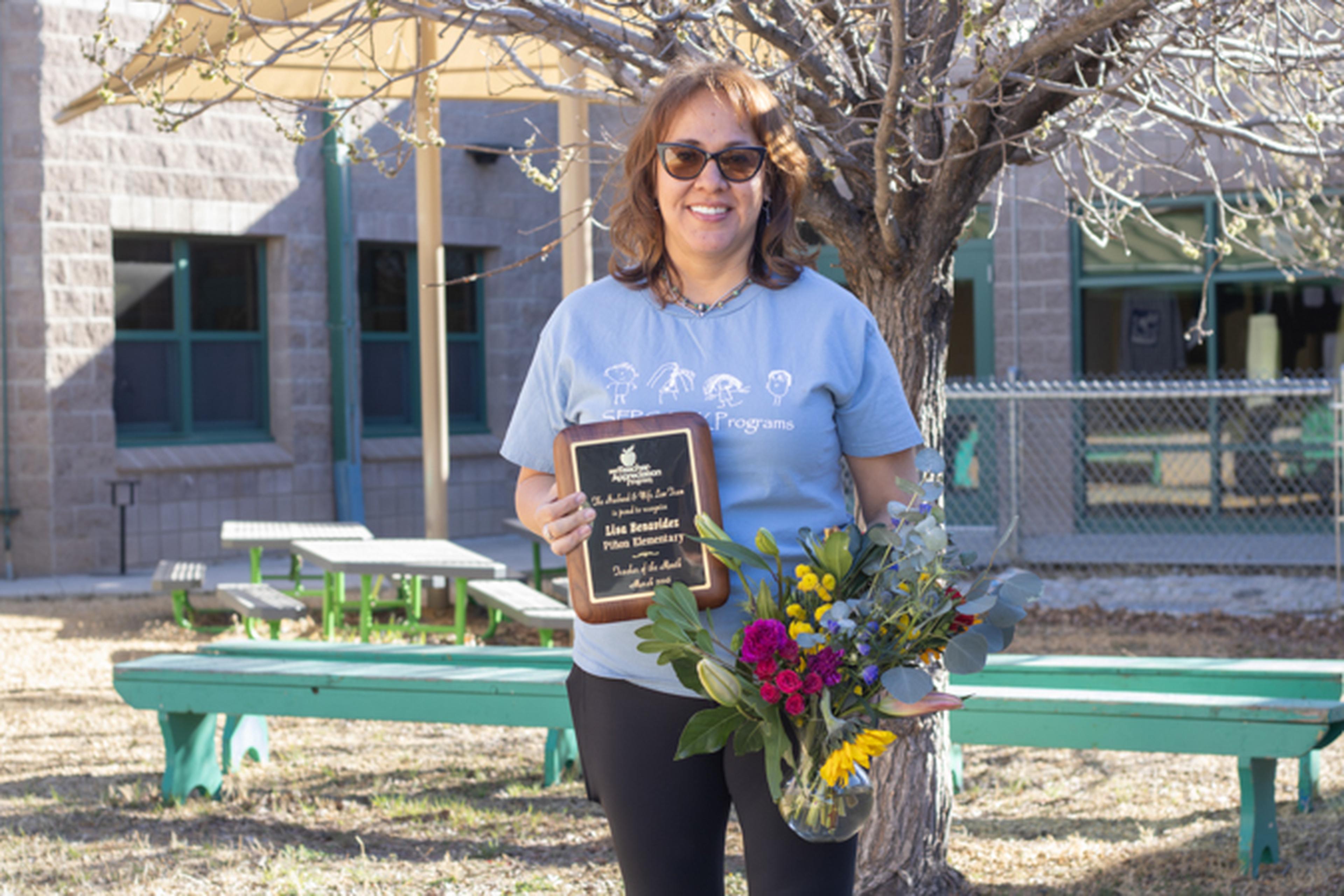 Ms. B, holds a plaque from The Husband & Wife Law Team and a bouquet of flowers, standing outside by a tree and benches.