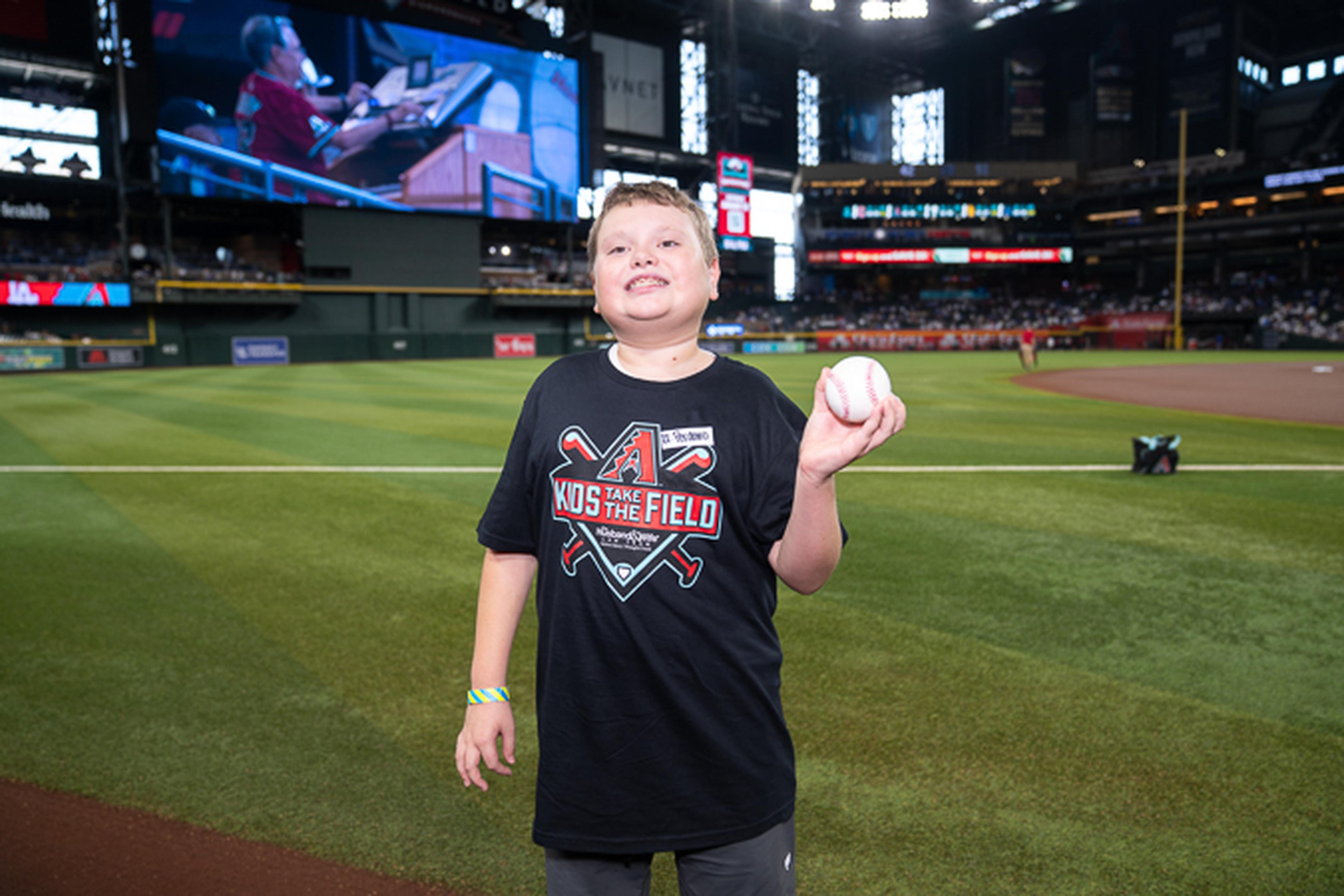 A young Kids Take the Field winner proudly holds a baseball on Chase Field, enjoying a memorable experience in front of a crowd of Arizona Diamondbacks fans.