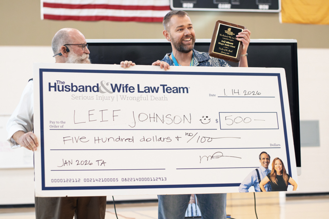 Mr. Johnson holding a large check and a plaque. The check is for $500, awarded by "The Husband & Wife Law Team" to Leif Johnson.