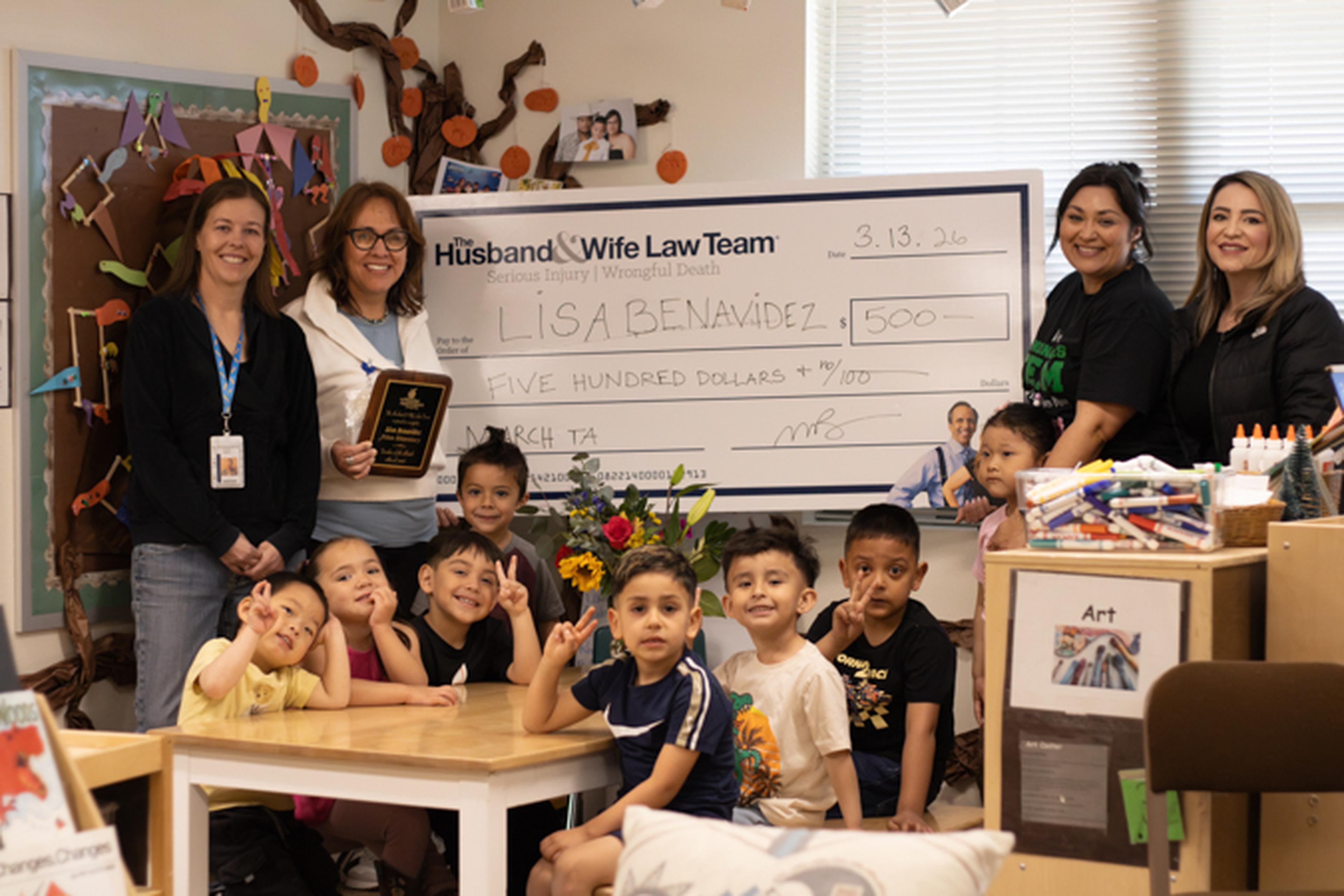 A group of teachers and young children from Piñon Elementary School pose with a large check for $500. A plaque and flowers are also present in the classroom setting.