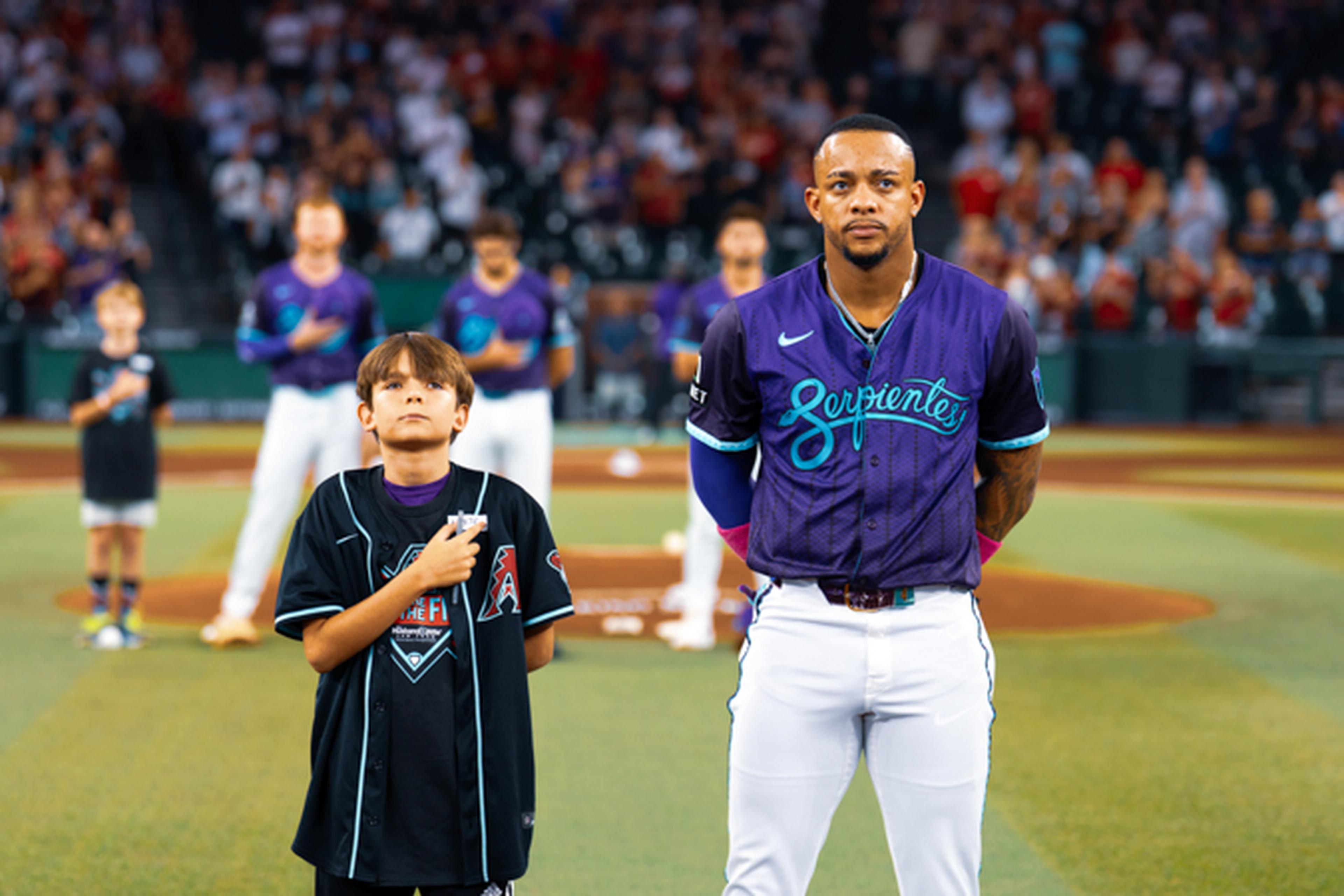 A baseball player and a child stand for the national anthem on a field, with other players and spectators in the background.