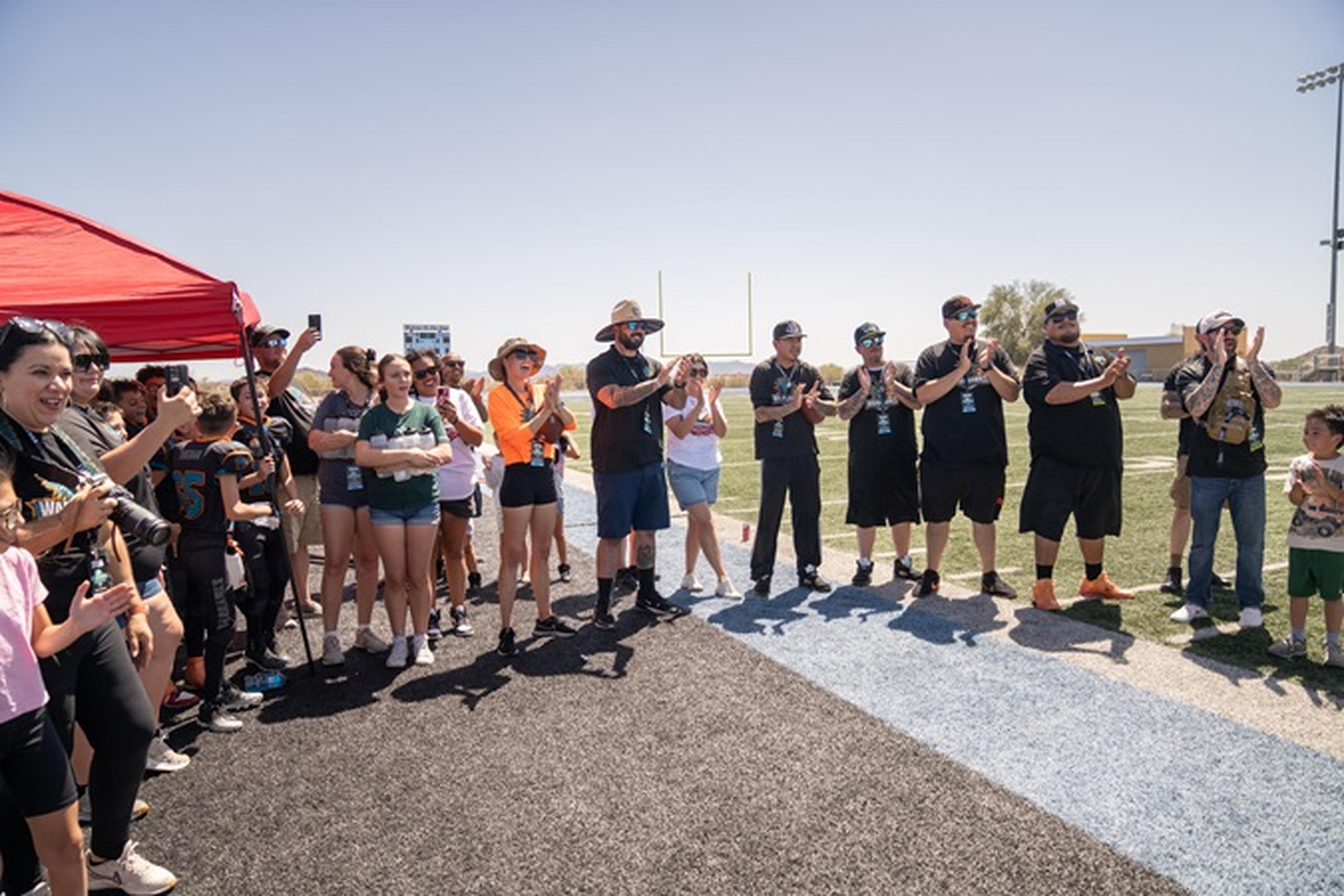 Arizona Youth Football League members gather on the field beneath a red tent, clapping and cheering under the bright blue Arizona sky.