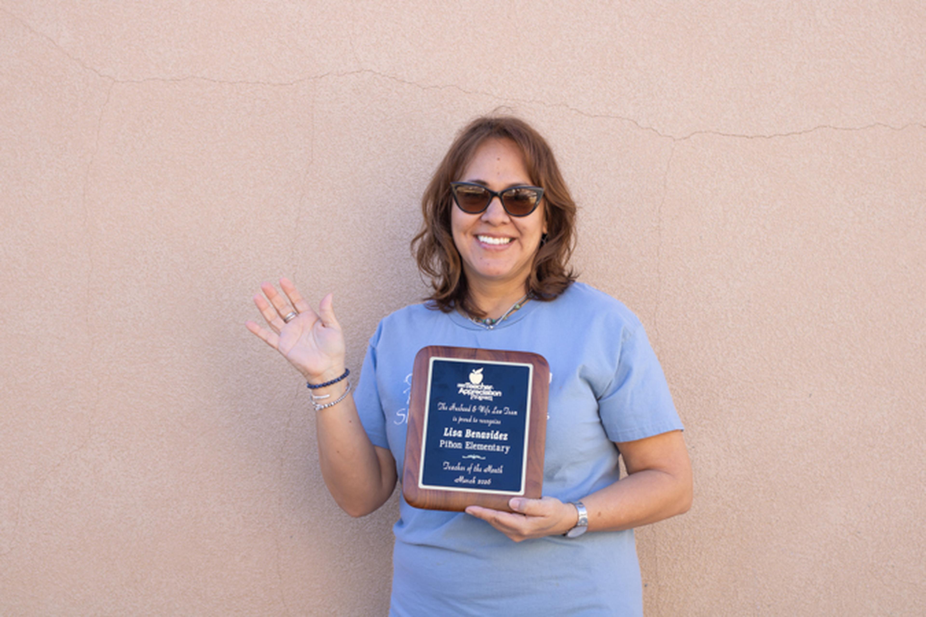 Ms. B, in sunglasses holds her Teacher Appreciation plaque and waves while standing against a beige wall.