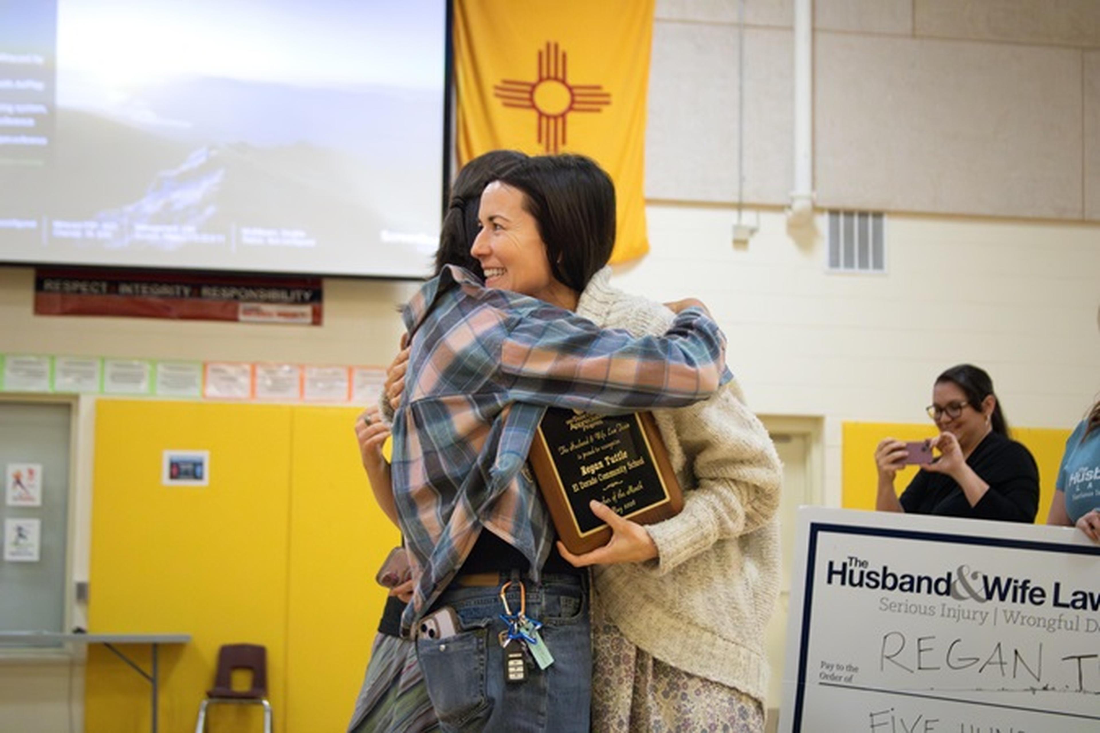 Two people hugging, one holding a plaque. A large check is visible, and a flag hangs in the background. Others are watching, smiling.