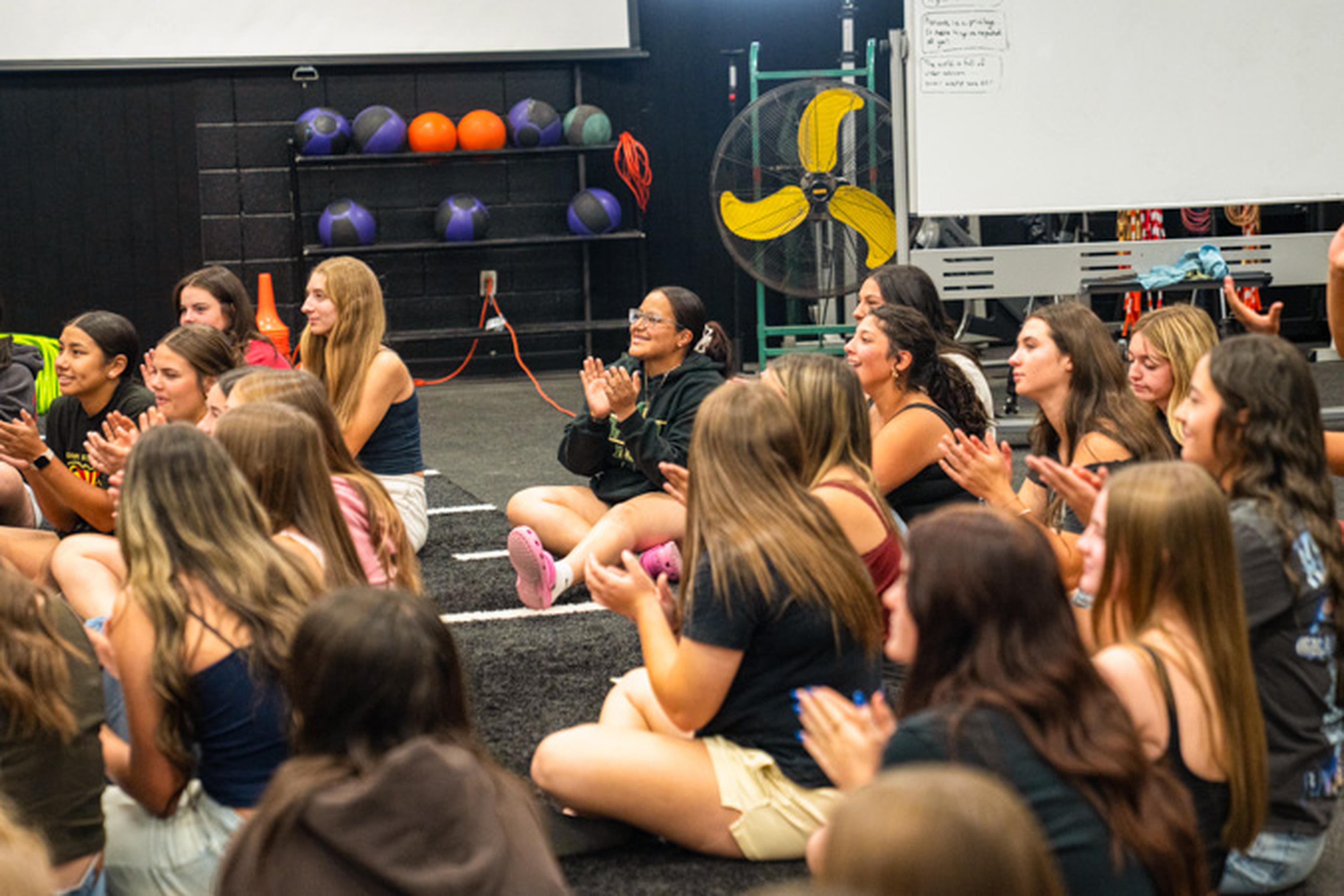 Basha athletes celebrate from the gym floor, clapping proudly for Coach Bright as workout gear and a large fan set the scene behind them.