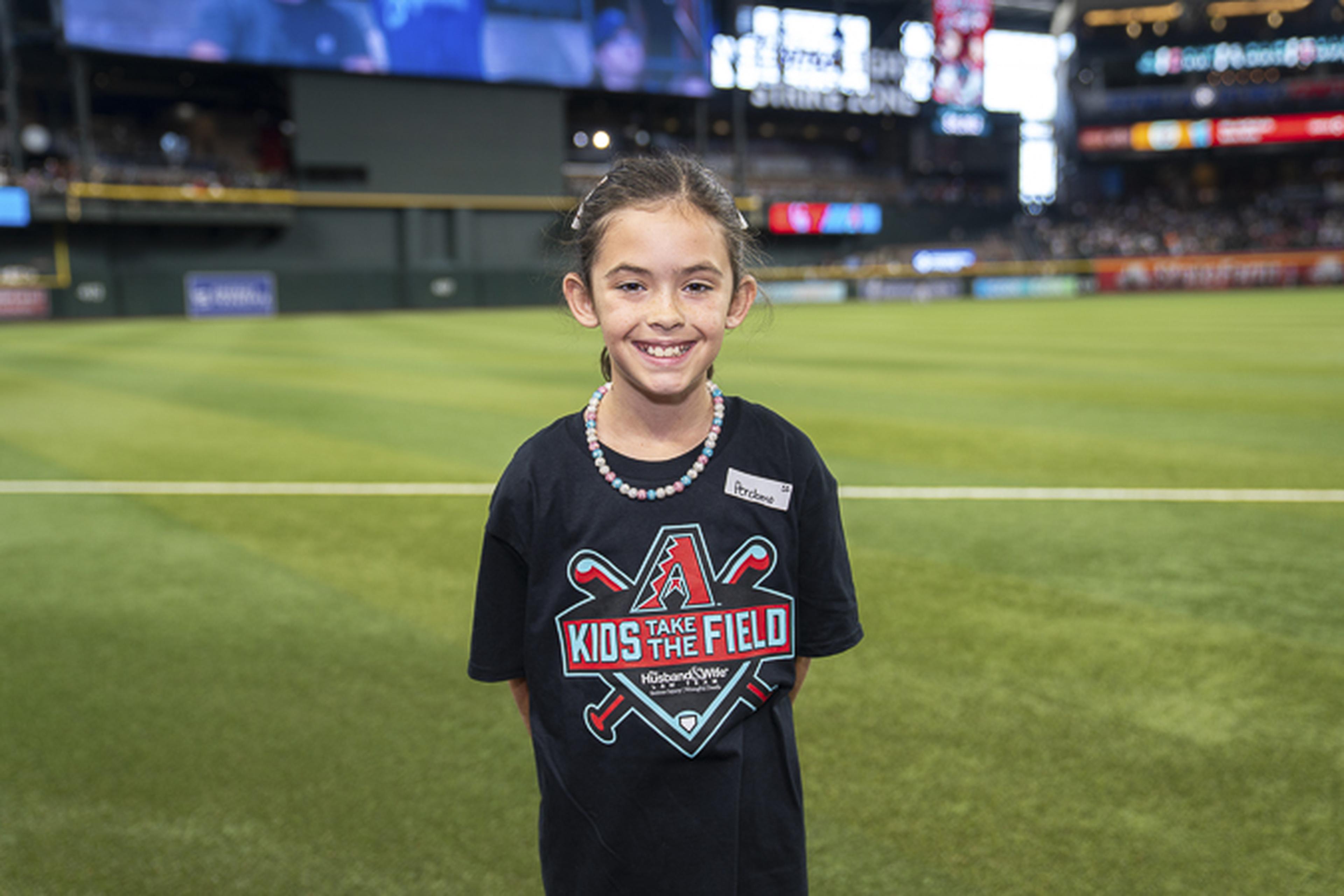 A smiling girl in a “Kids Take the Field” shirt stands on the baseball field with the stadium rising behind her.