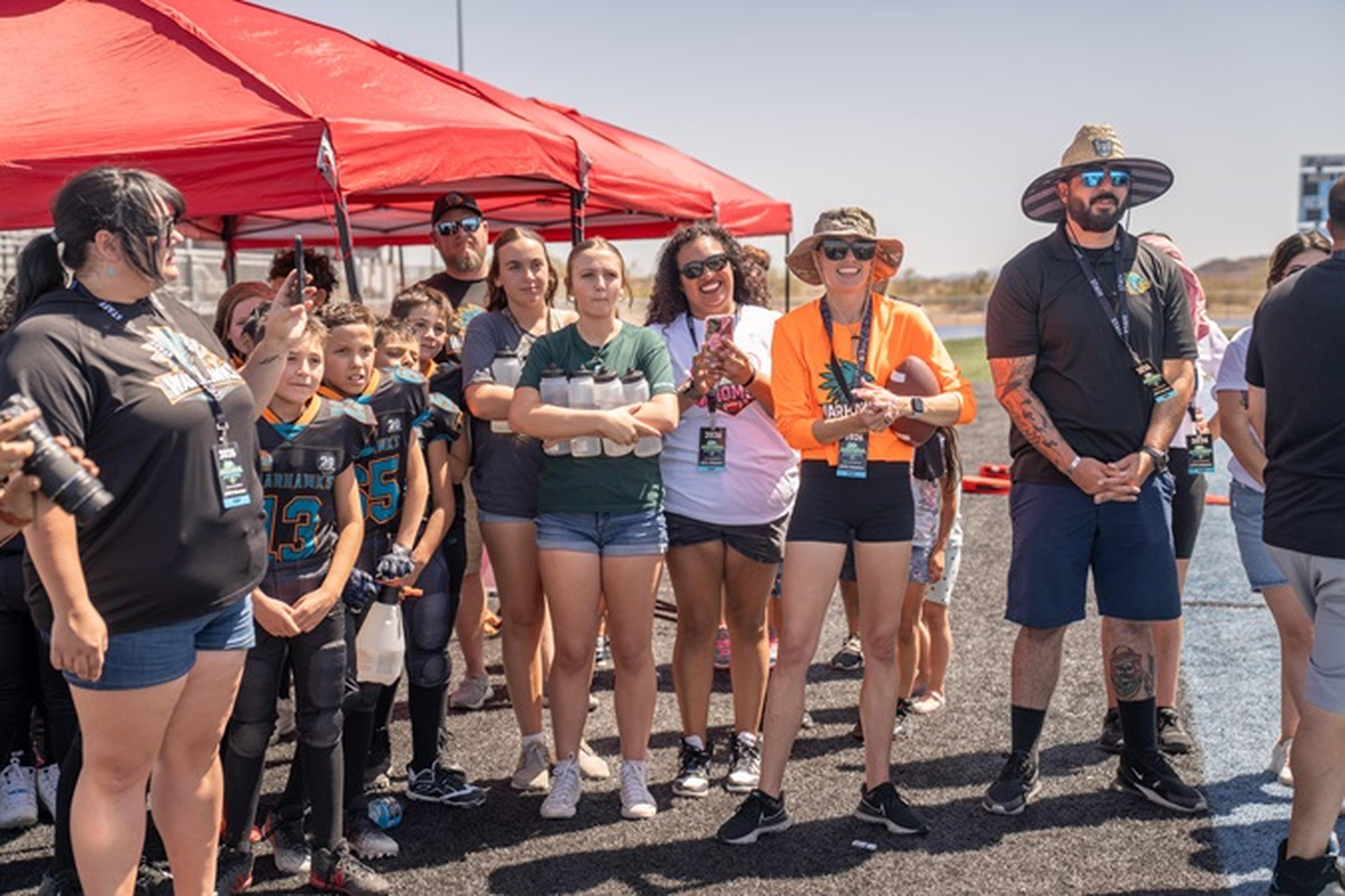 Arizona Youth Football League supporters, including kids in matching jerseys, gather under a red canopy on a sunny day as they watch practice.