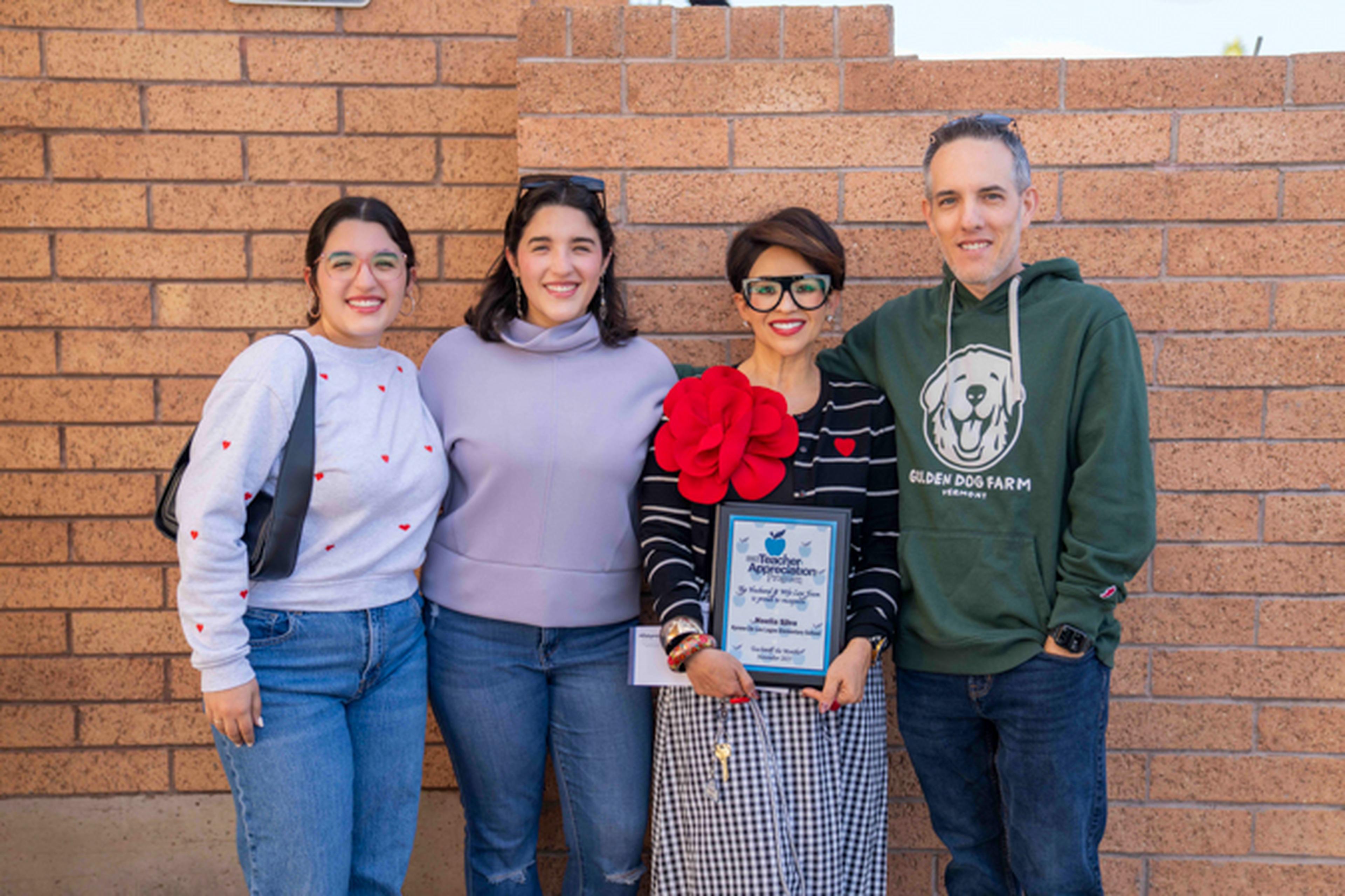 three women and one man stand in front of a school wall holding a plaque for Teacher Appreciation